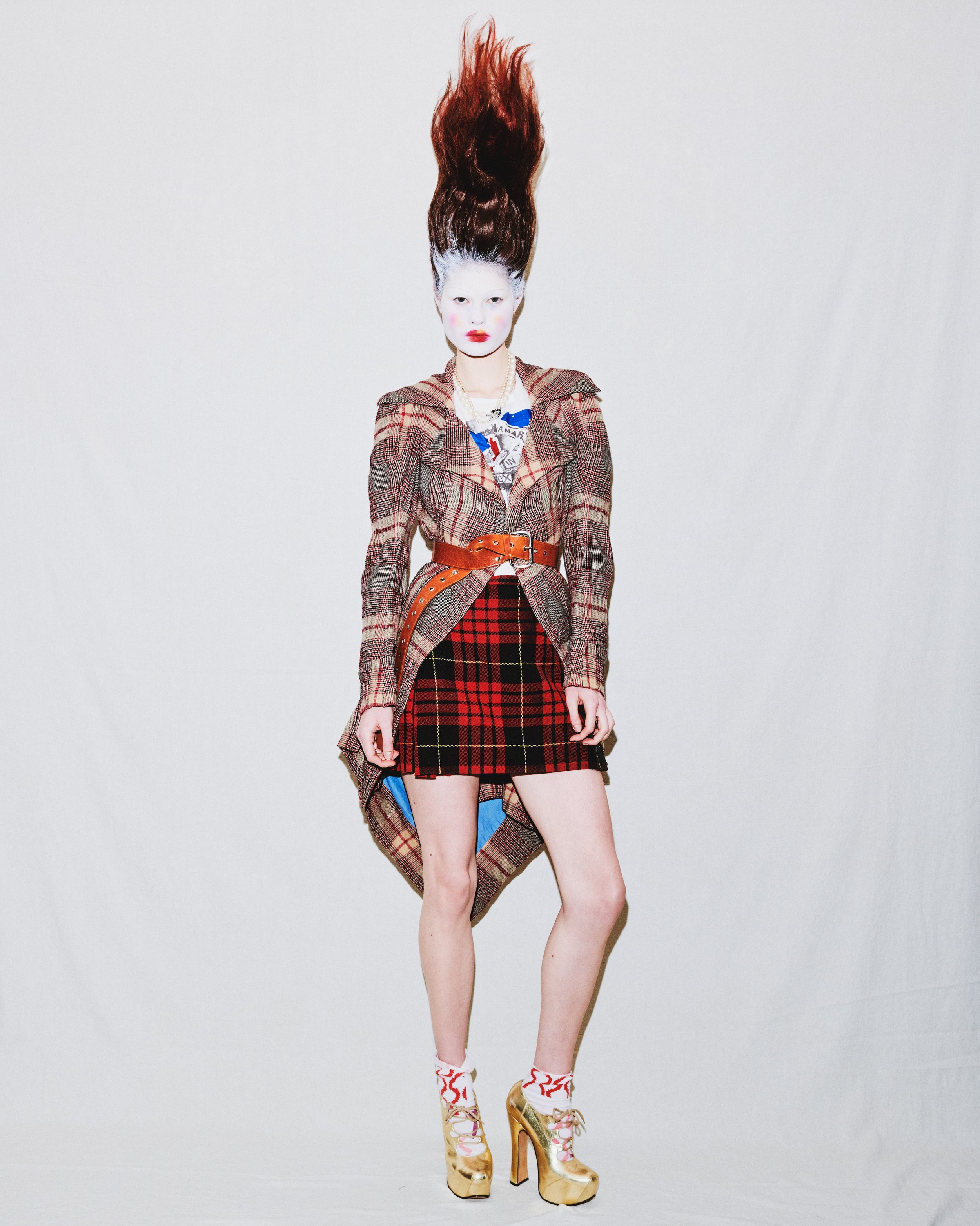 A woman with platinum blonde hair and face paint, wearing a plaid jacket, a red and black plaid skirt, gold high-heeled shoes, patterned socks, and layered necklaces, standing against a plain white background.