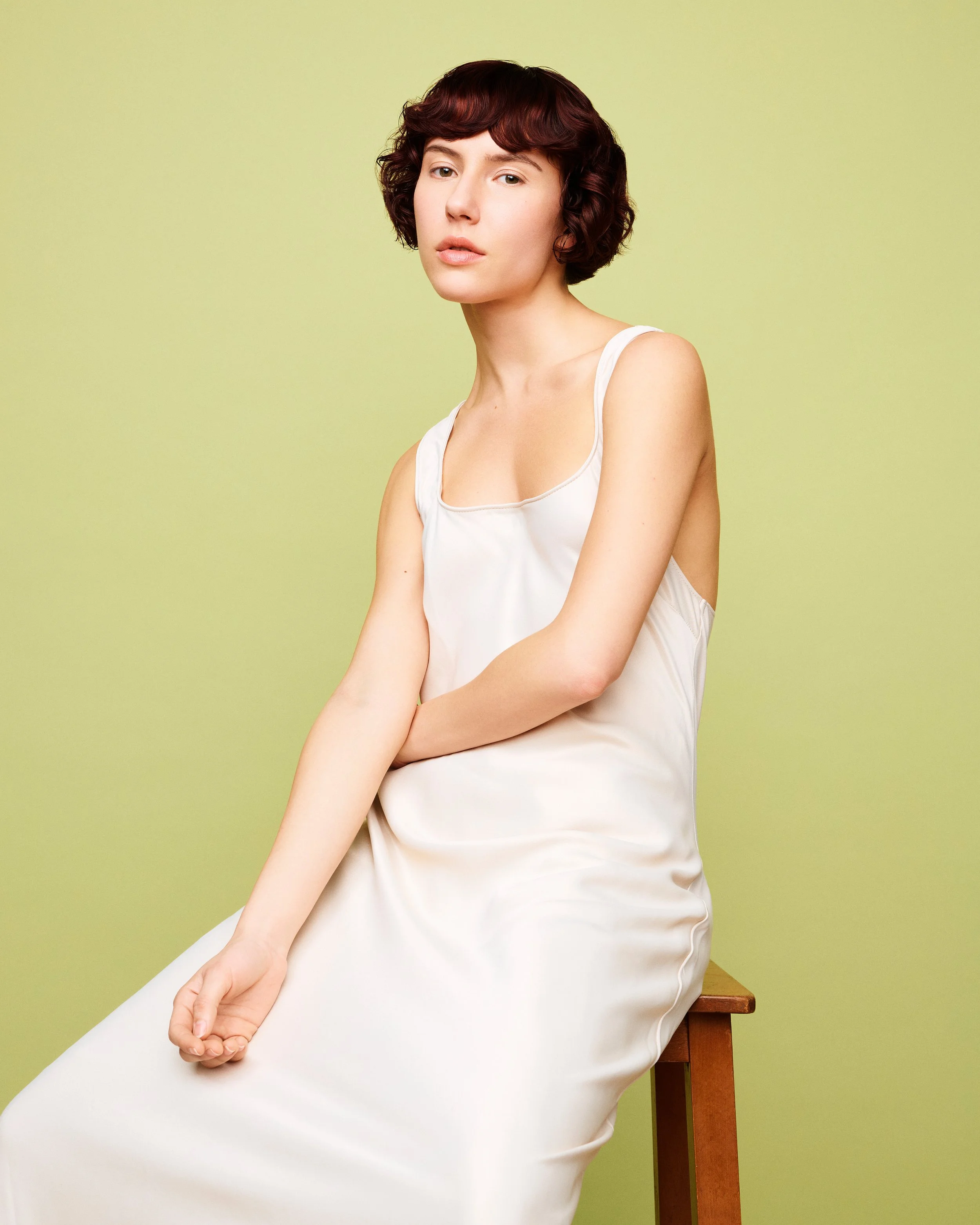 A woman with short, wavy, auburn hair wearing a white sleeveless top sitting on a wooden stool against a light green wall.