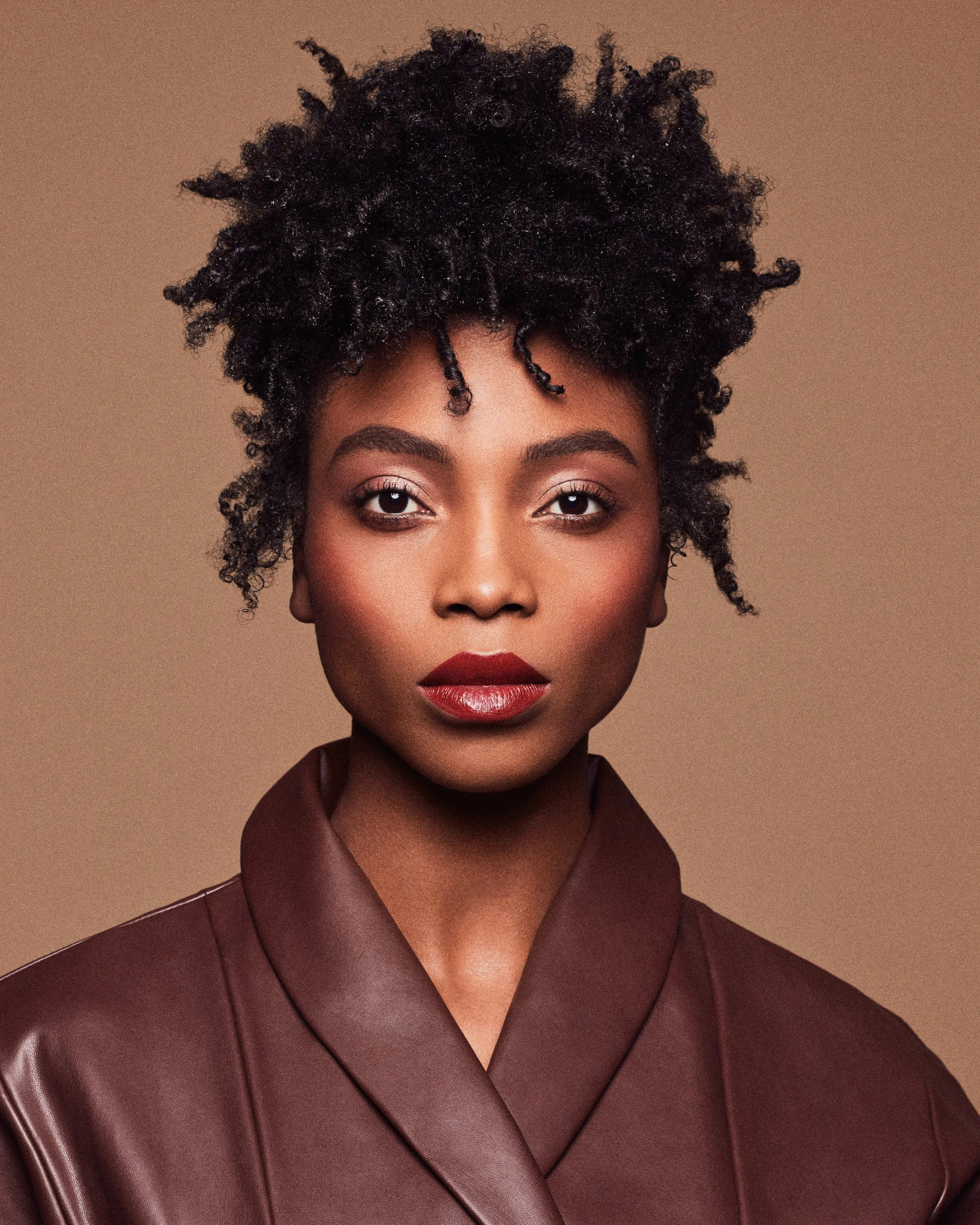 Close-up of a woman with natural hair in a messy curly updo, wearing a burgundy blazer, against a neutral background.