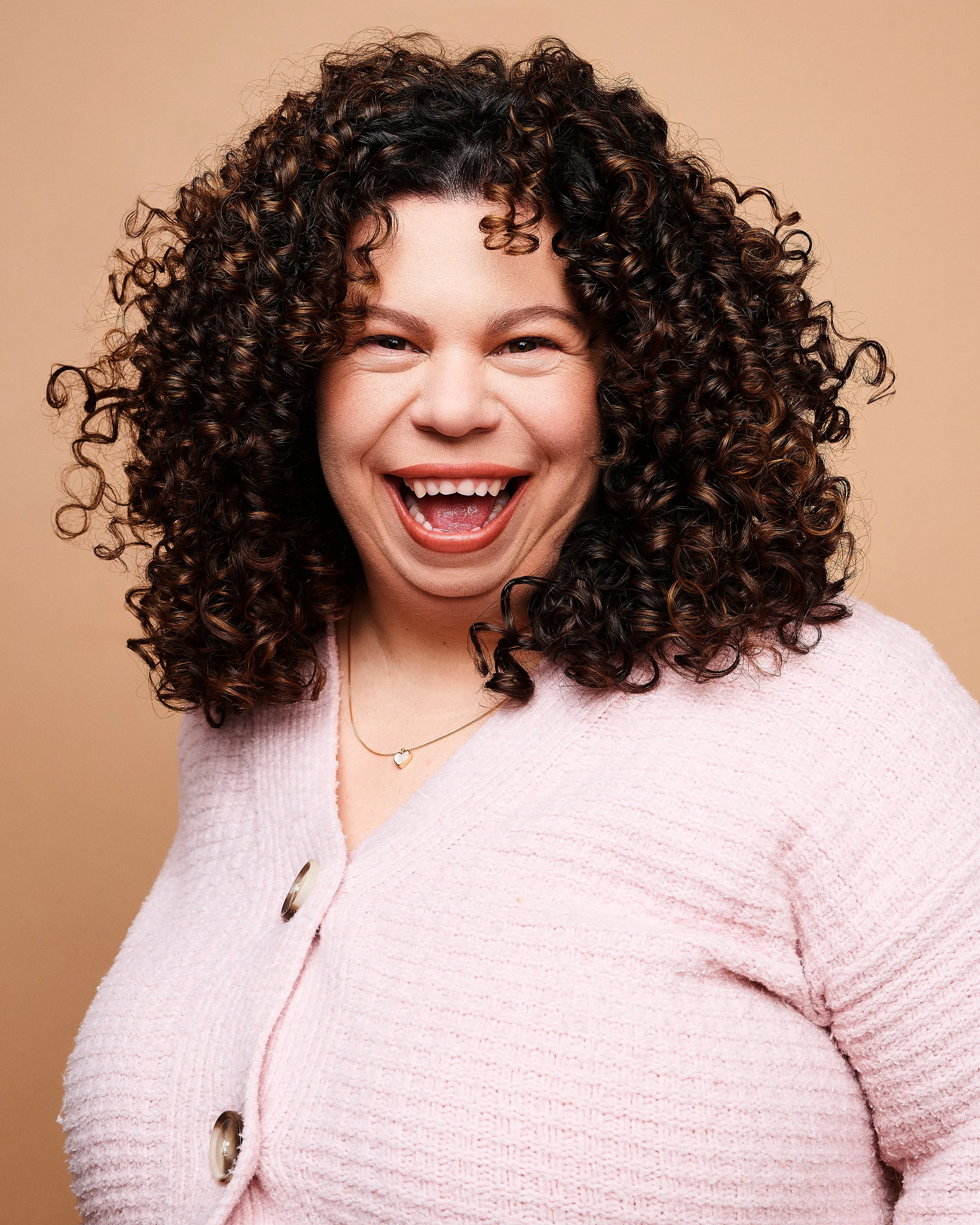 Close-up of a woman with curly hair, smiling and laughing, wearing a pink textured sweater and a heart-shaped necklace, against a neutral background.