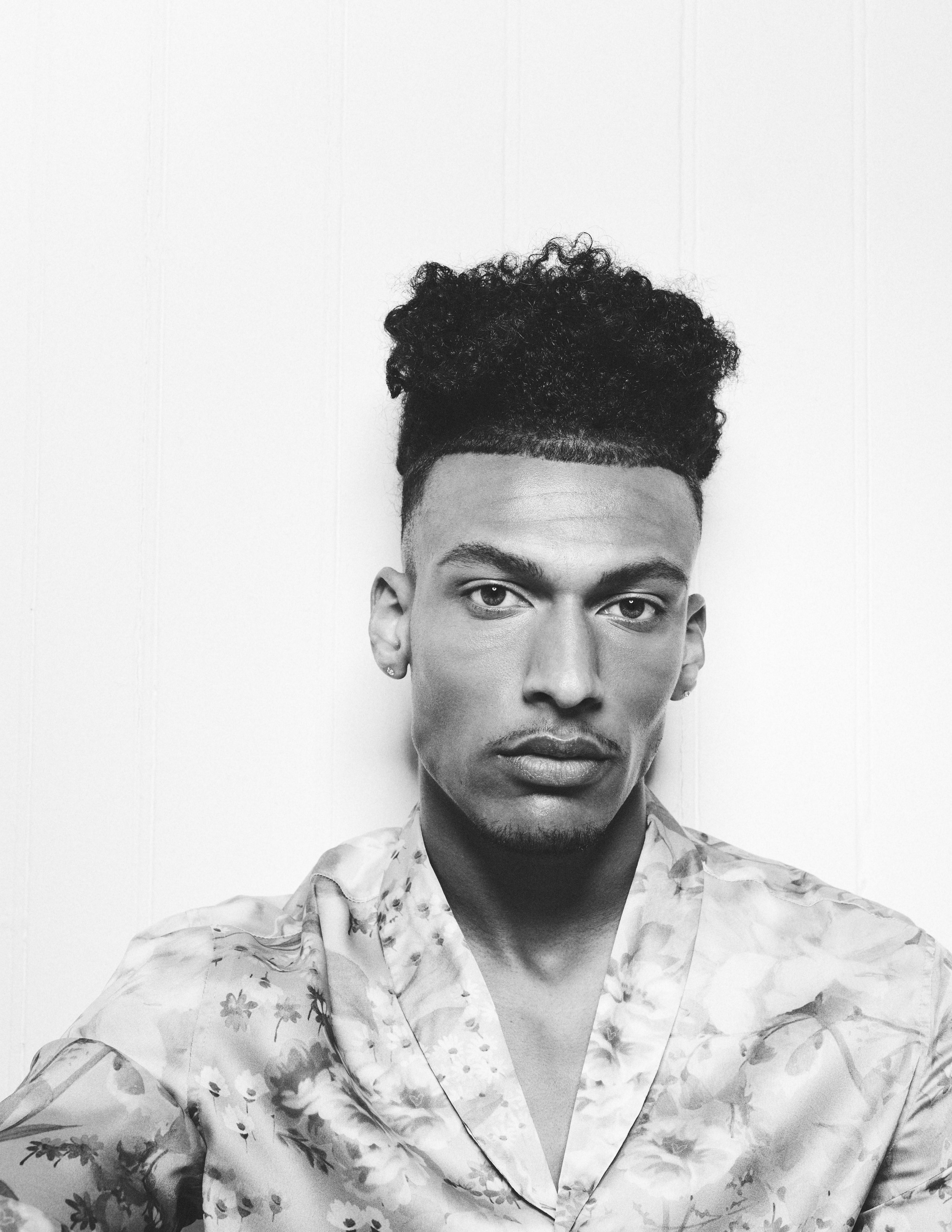 Black and white portrait of a young man with styled curly hair, wearing a floral patterned shirt, against a plain background.
