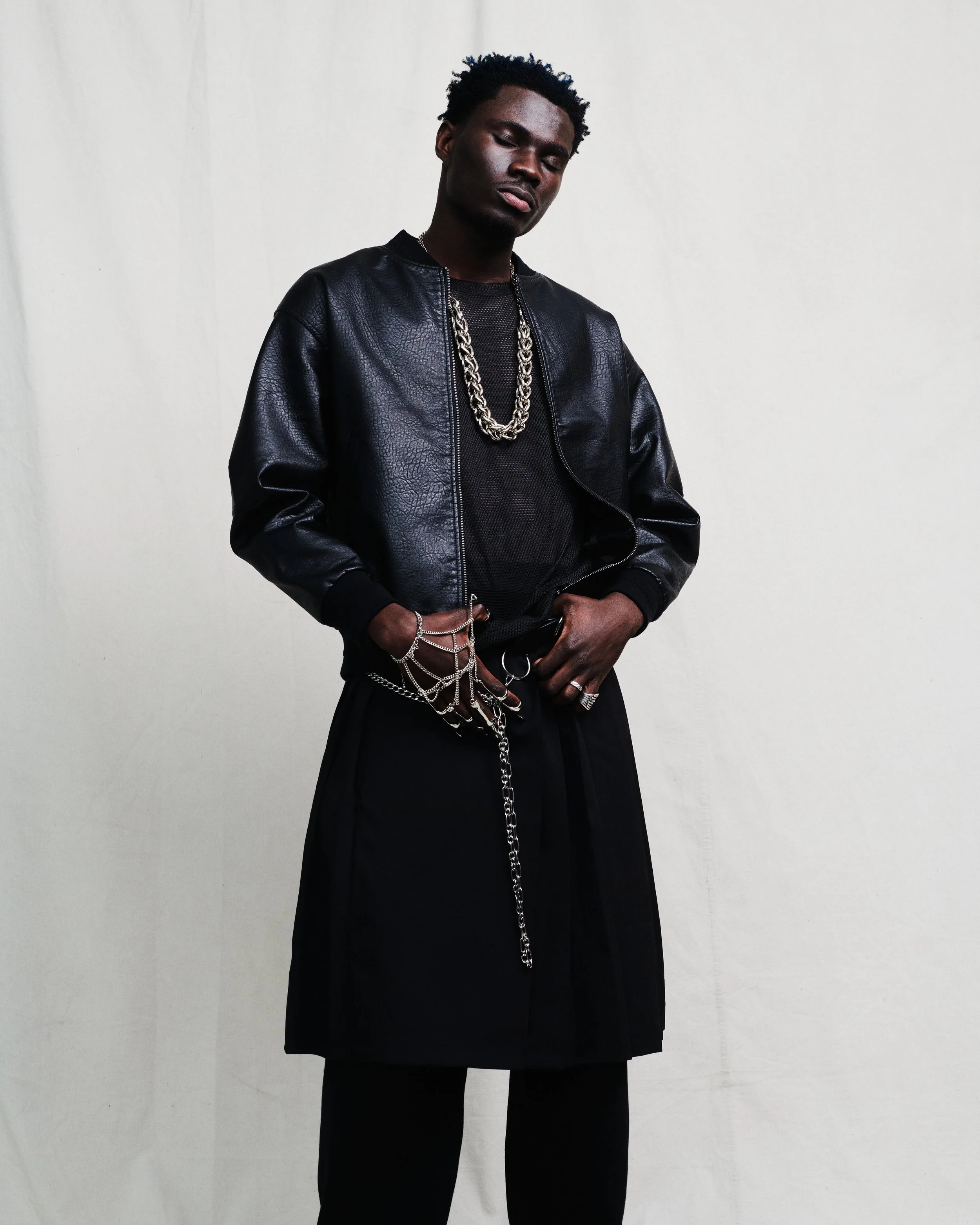 A young man with dark skin, short curly hair, wearing a black leather jacket, a black mesh shirt, layered silver necklaces, multiple silver rings, and silver chains hanging from his fingers, standing against a plain white wall.