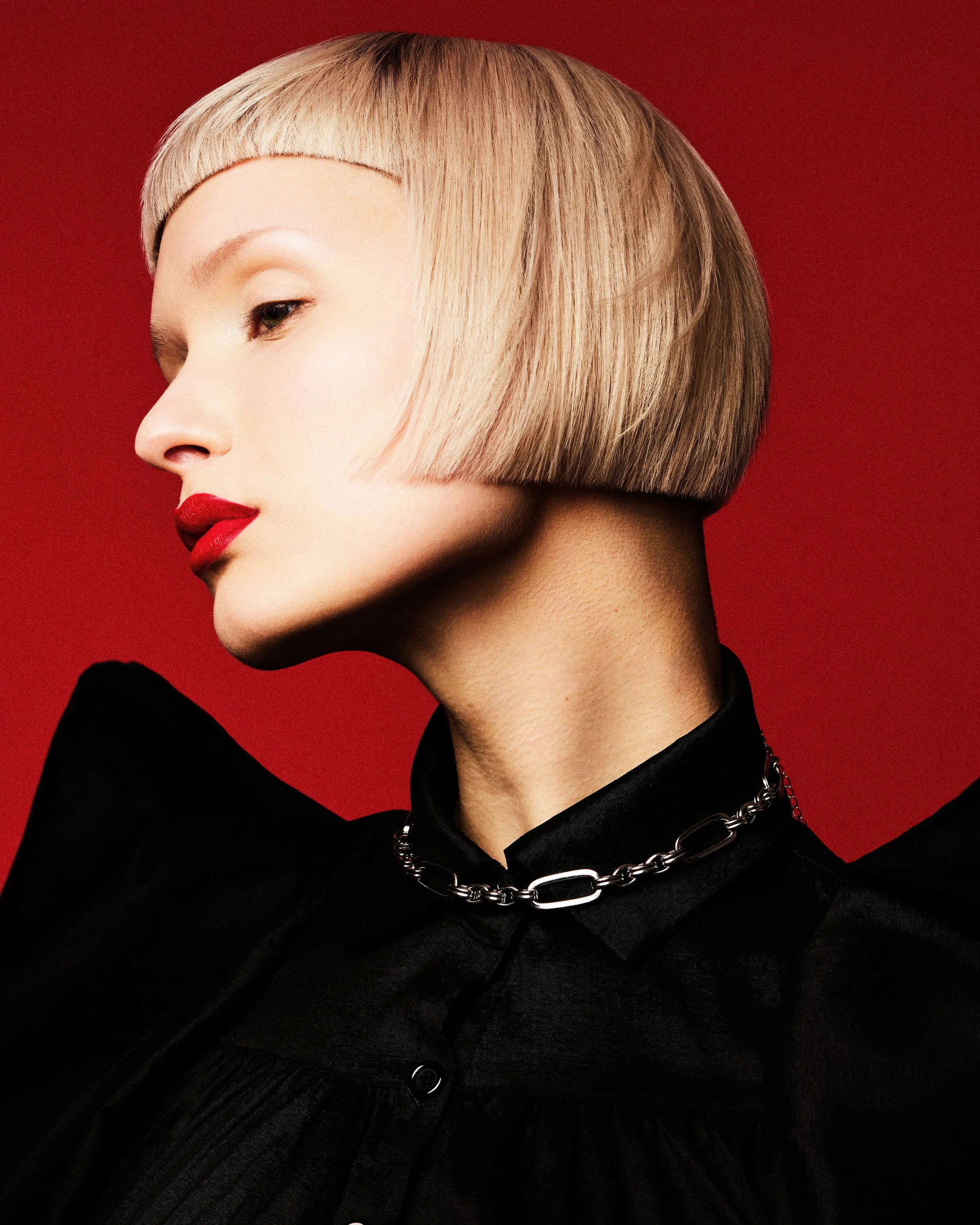 A woman with light blonde hair styled in a straight bob with blunt bangs, wearing red lipstick, a black outfit, and a silver chain necklace, against a red background.
