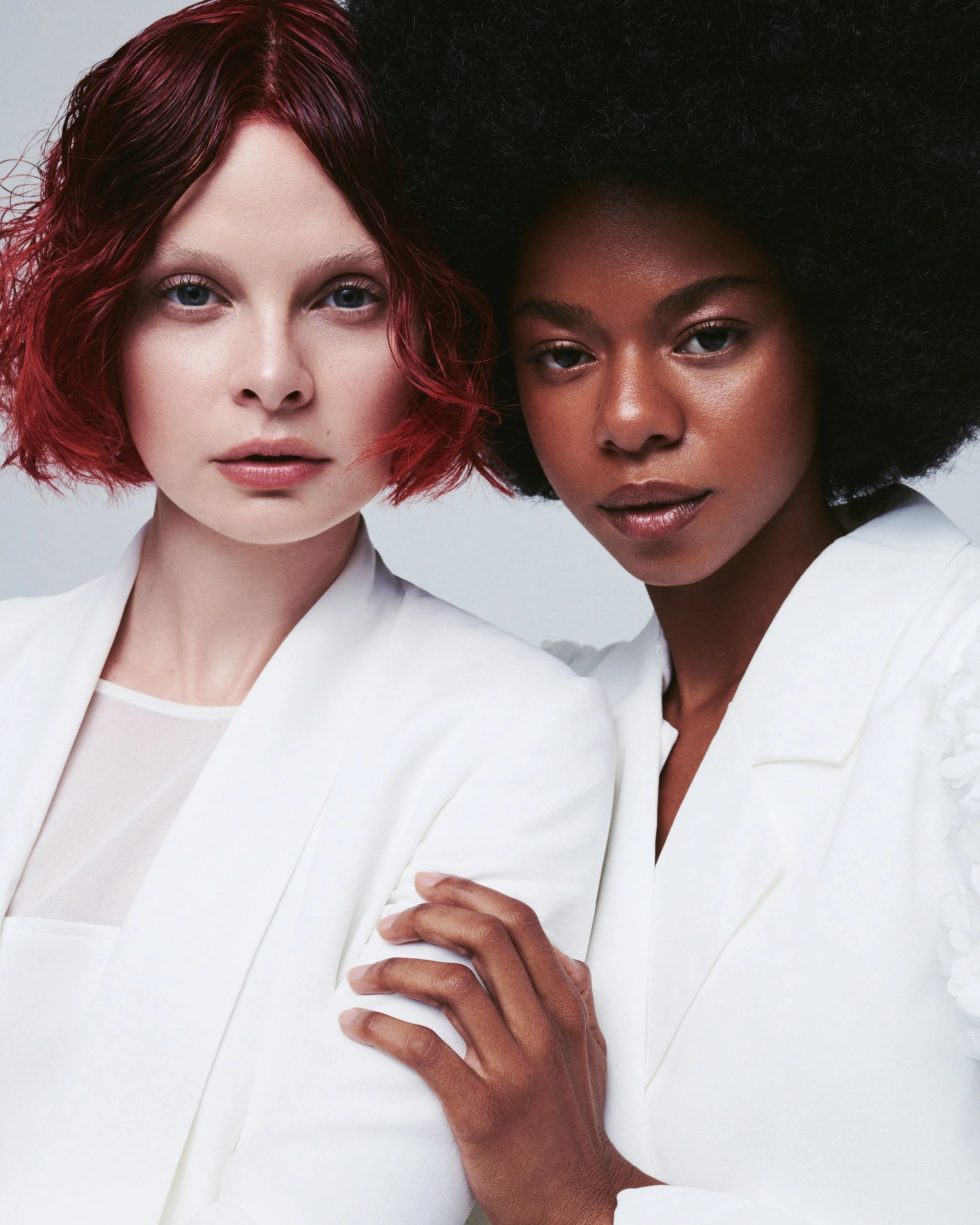 Close-up of two women with different ethnicities posing together wearing white clothing, one with red hair and the other with black curly hair.