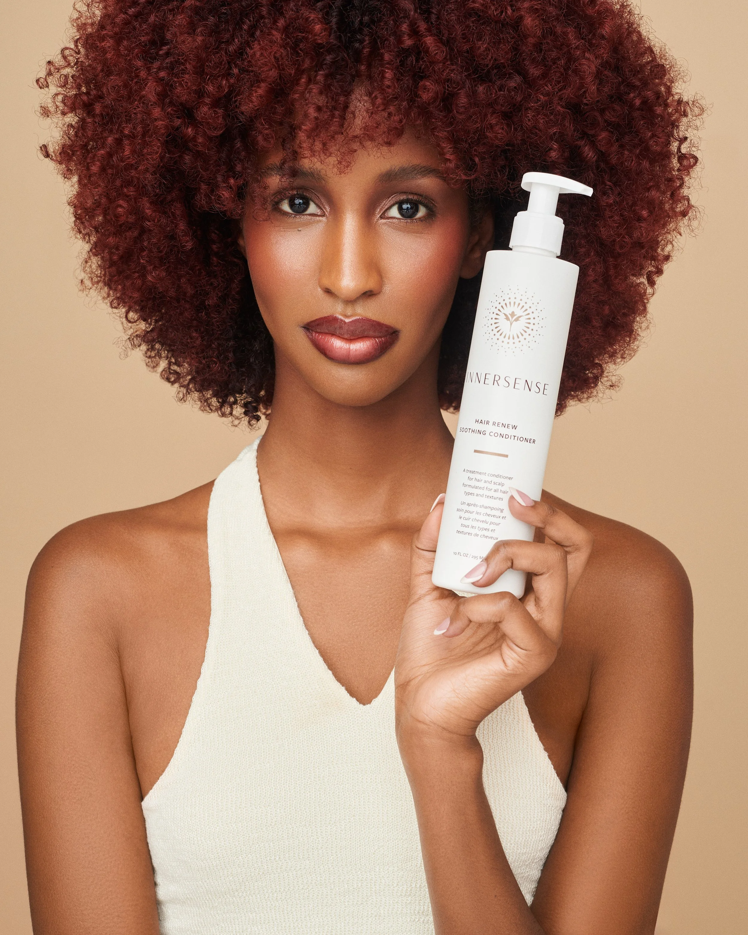 A woman with curly red hair holding a white bottle of Innersense hair conditioner close to her face. She has smooth skin and is wearing a sleeveless, cream-colored top against a neutral background.