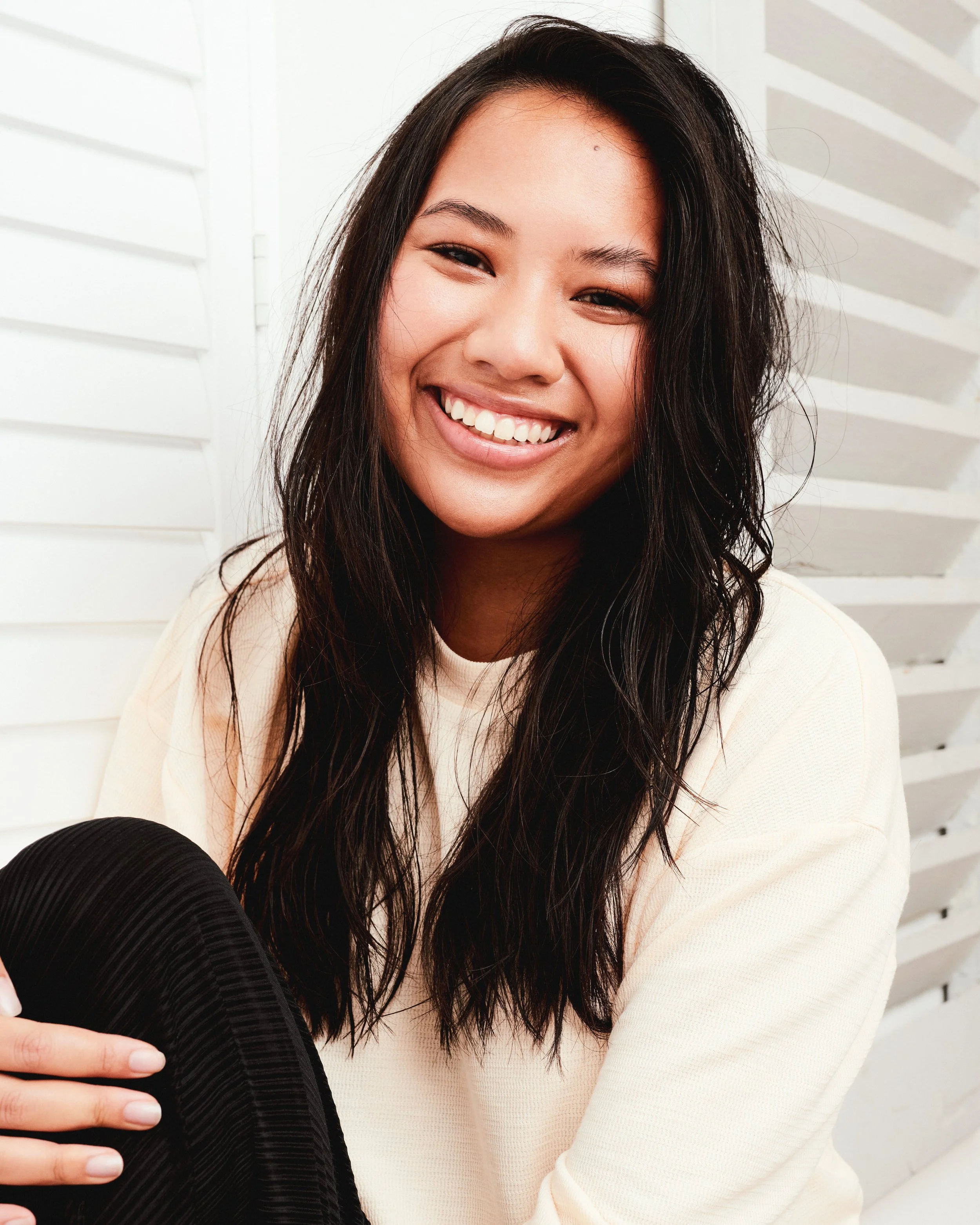 A young woman with long dark hair smiling and looking at the camera, wearing a light-colored sweater, sitting outdoors near a white louvered wall.