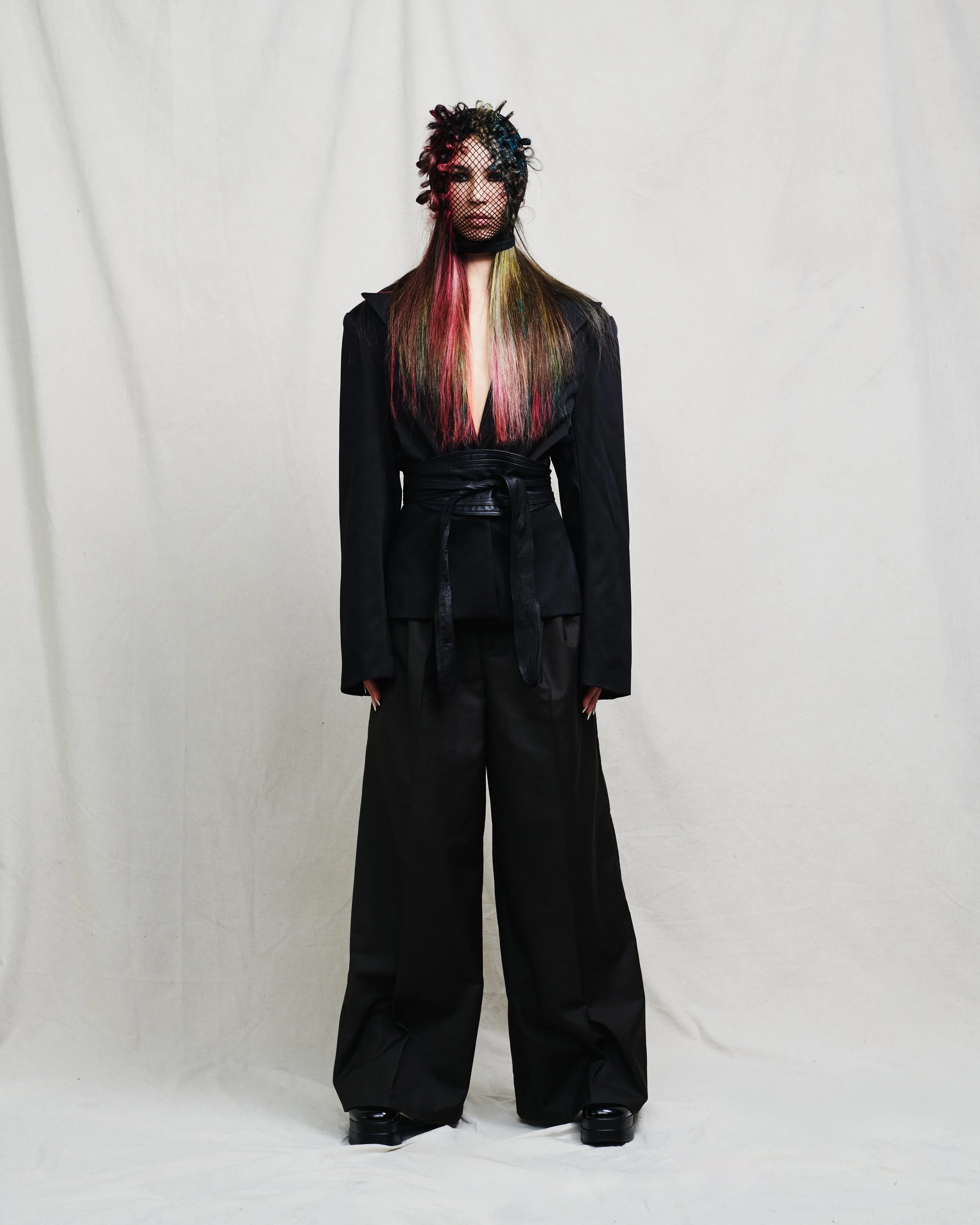 A person in black clothing wearing platform shoes, a fishnet mask, and with colorful hair against a plain white background.