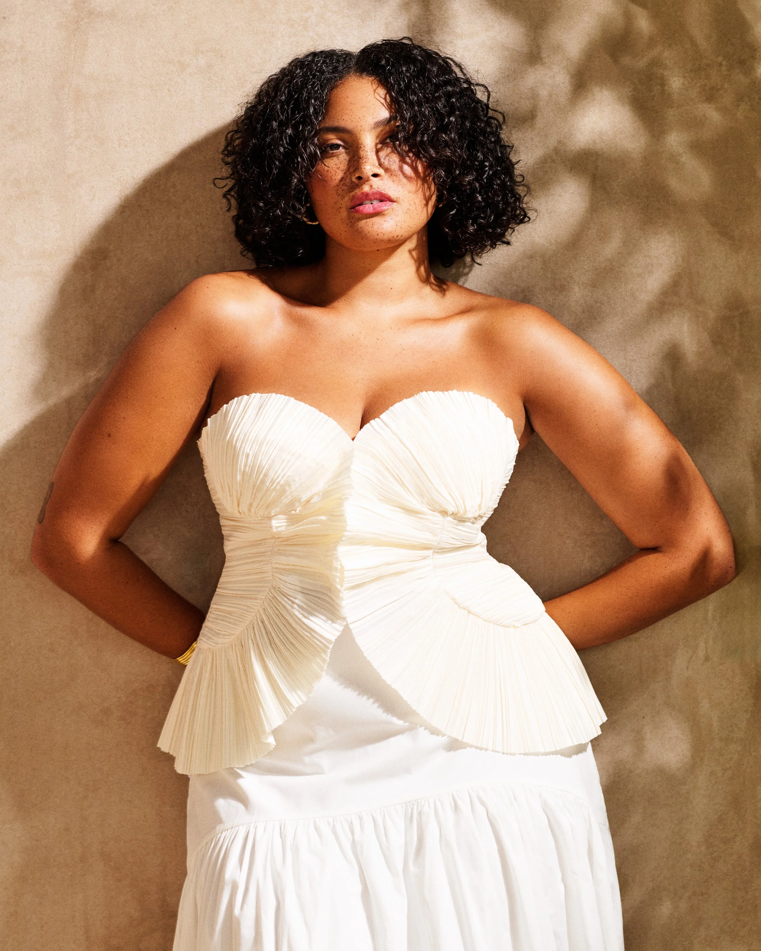 A woman with curly dark hair and freckles wearing a strapless white dress, lying on a beige surface with shadows.