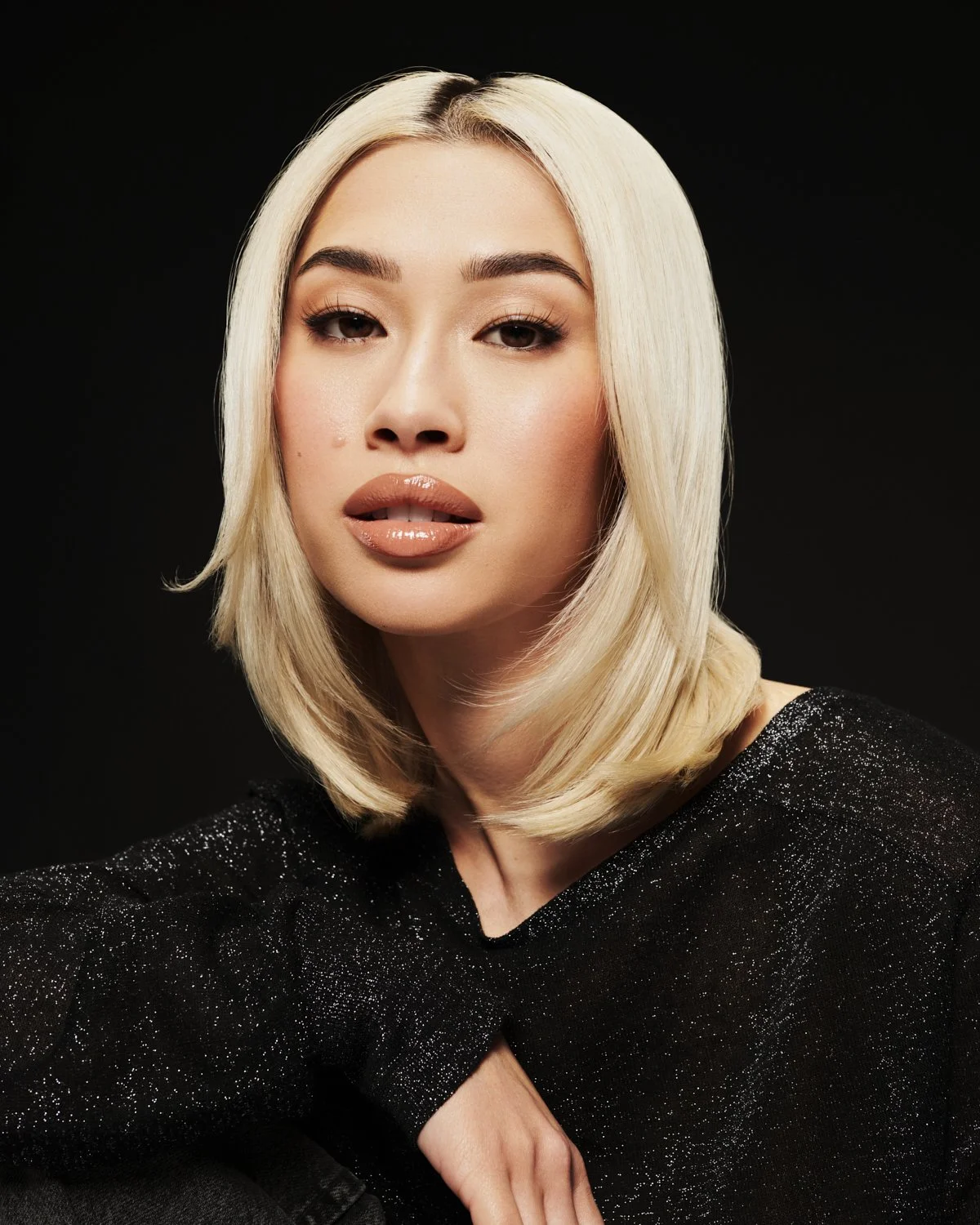 A woman with platinum blonde hair styled in a bob, wearing a black sparkly top, looking directly at the camera against a black background.