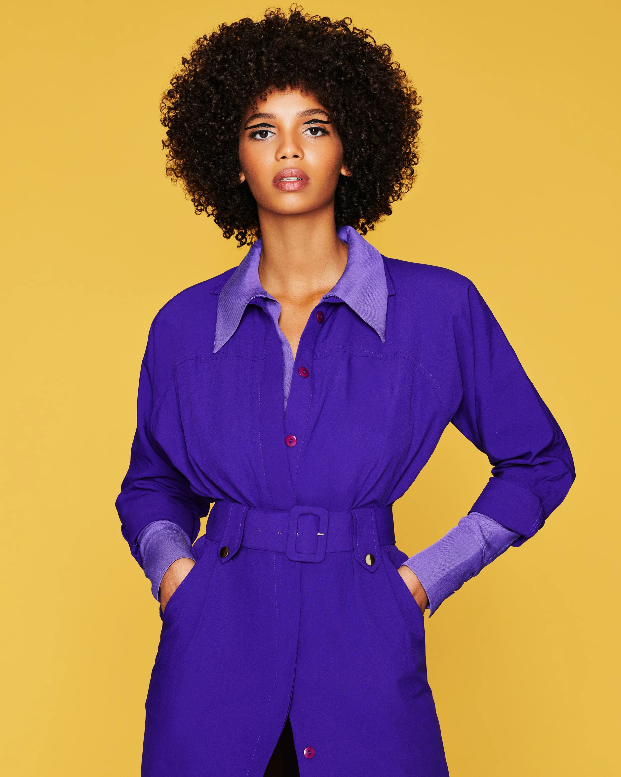 A woman with curly black hair wearing a purple jumpsuit with rolled-up sleeves against a yellow background.