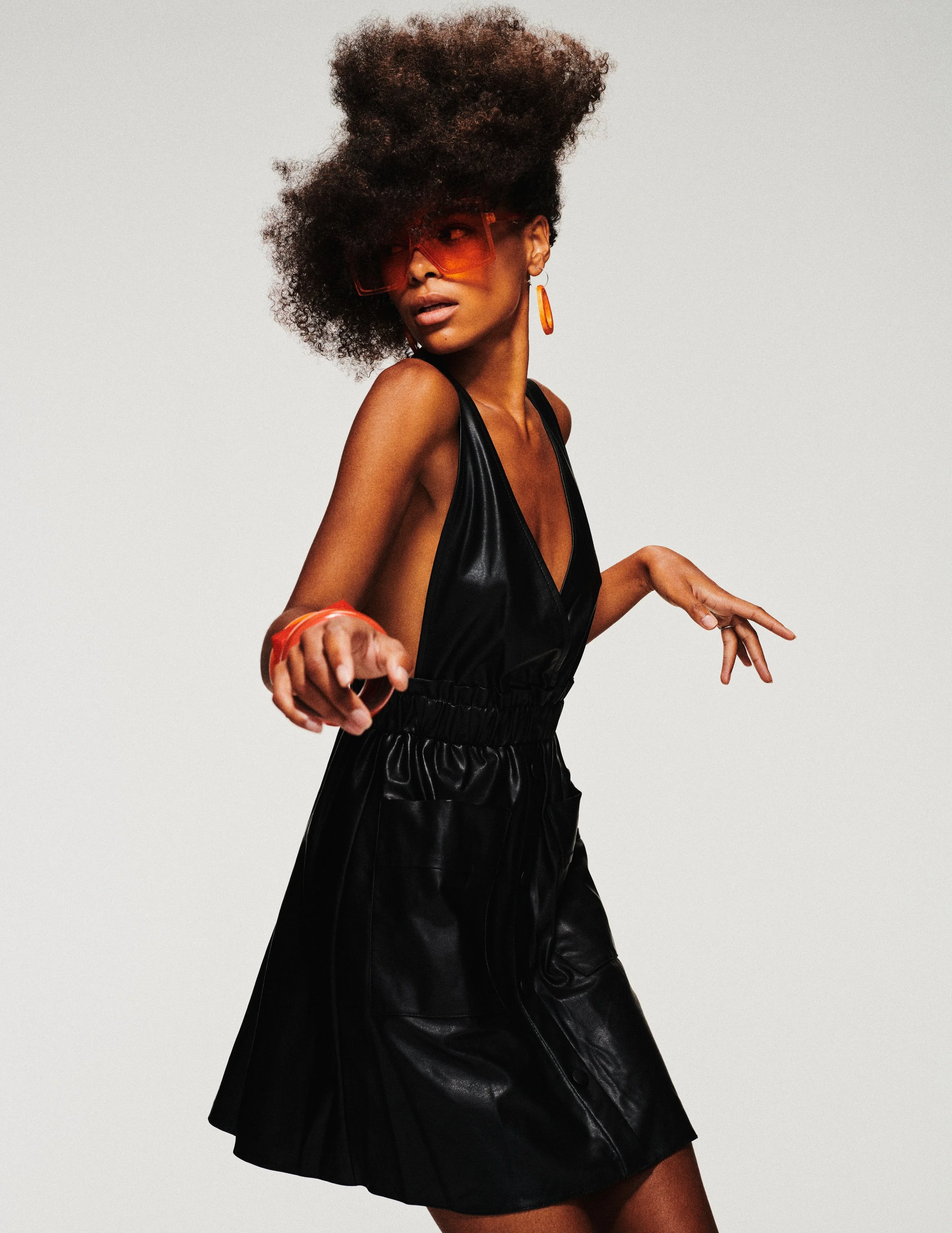 A woman with dark skin, curly hair styled in an updo, wearing large orange sunglasses, and dangling orange earrings. She is dressed in a black leather top and skirt, striking a pose against a plain white background.
