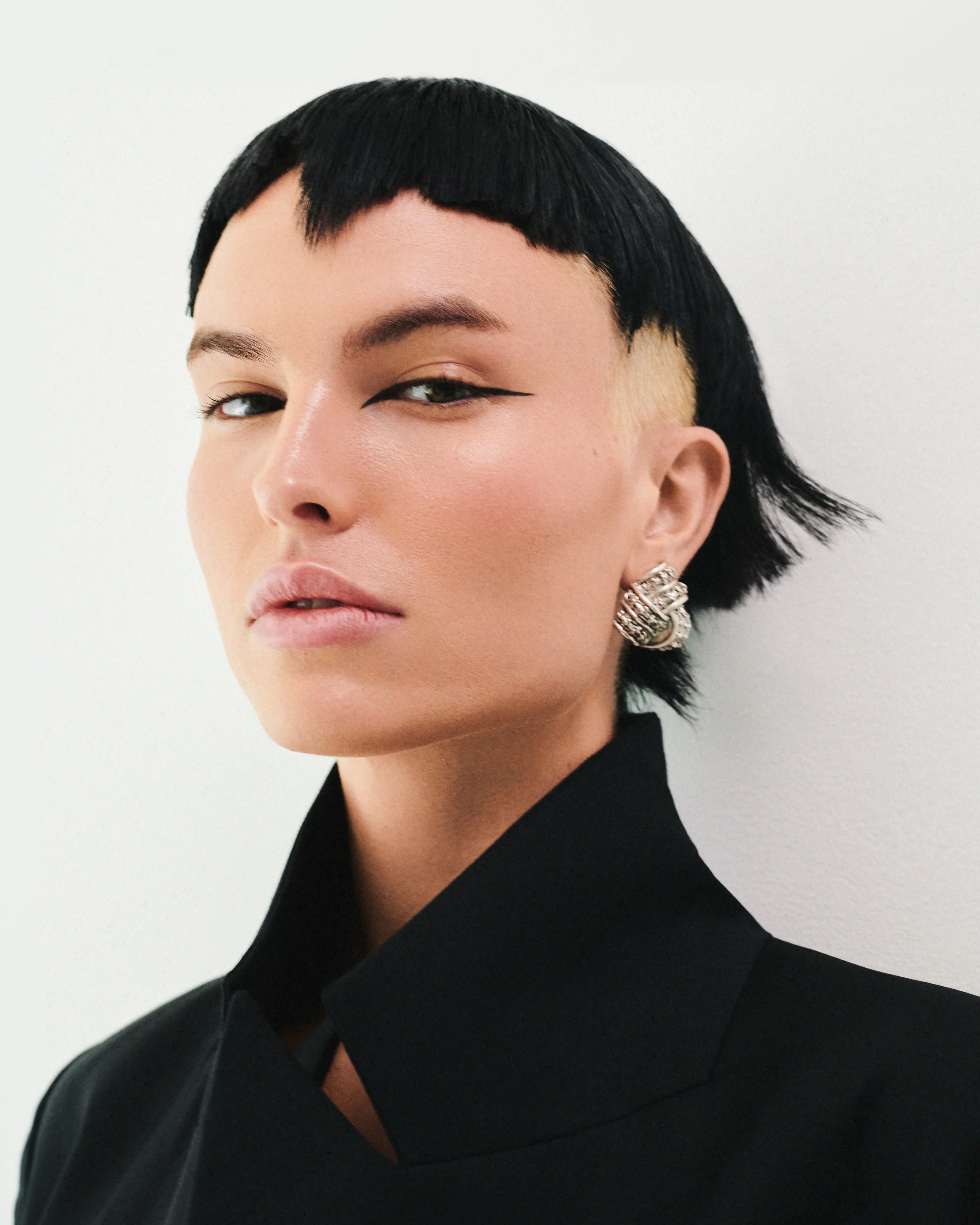 Close-up of a person with short black hair styled in a layered bob, wearing a black jacket and silver earrings, against a plain white background.