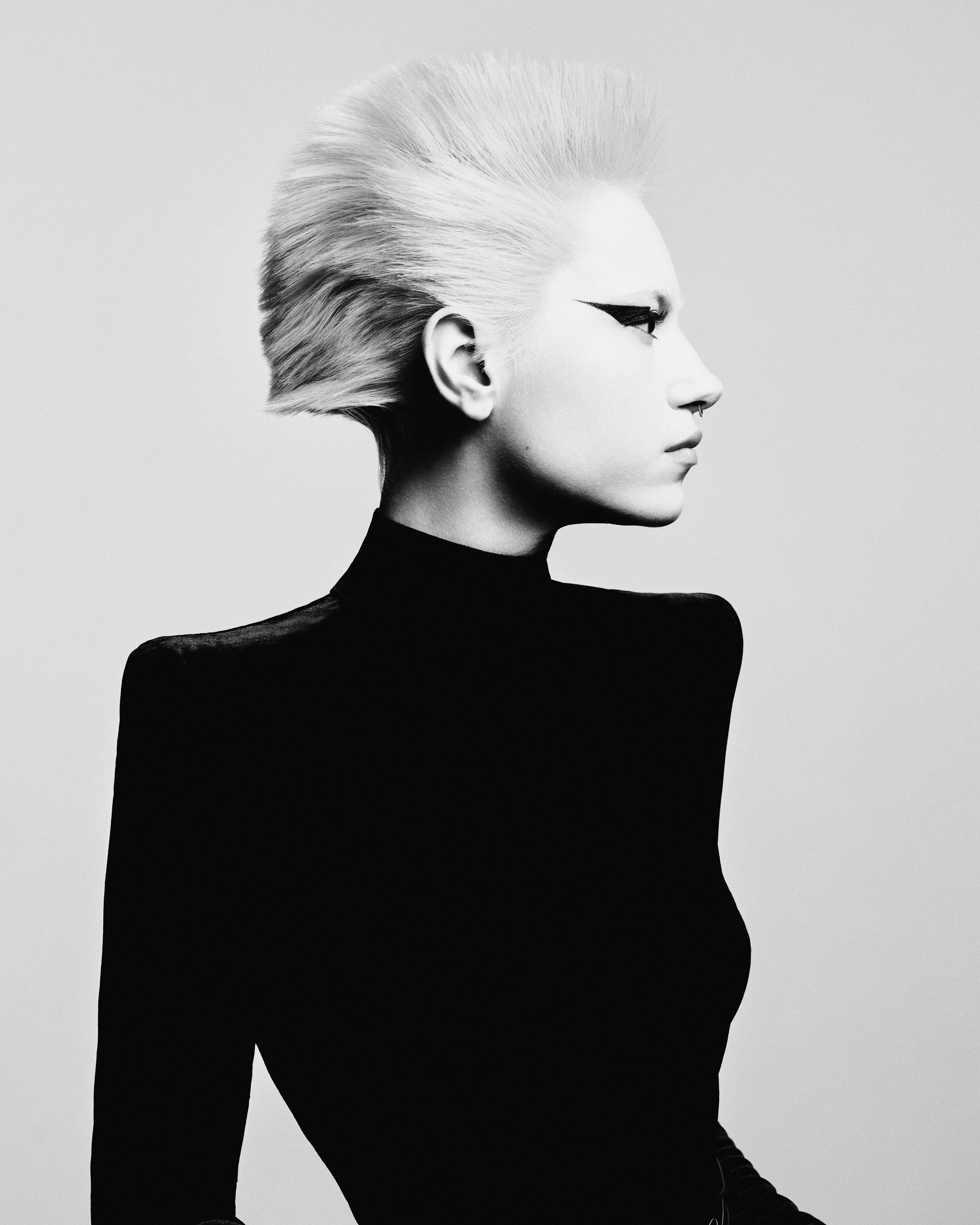 Black and white profile of a woman with platinum blonde hair styled in a pompadour, wearing a black turtleneck and bold black eyeliner.