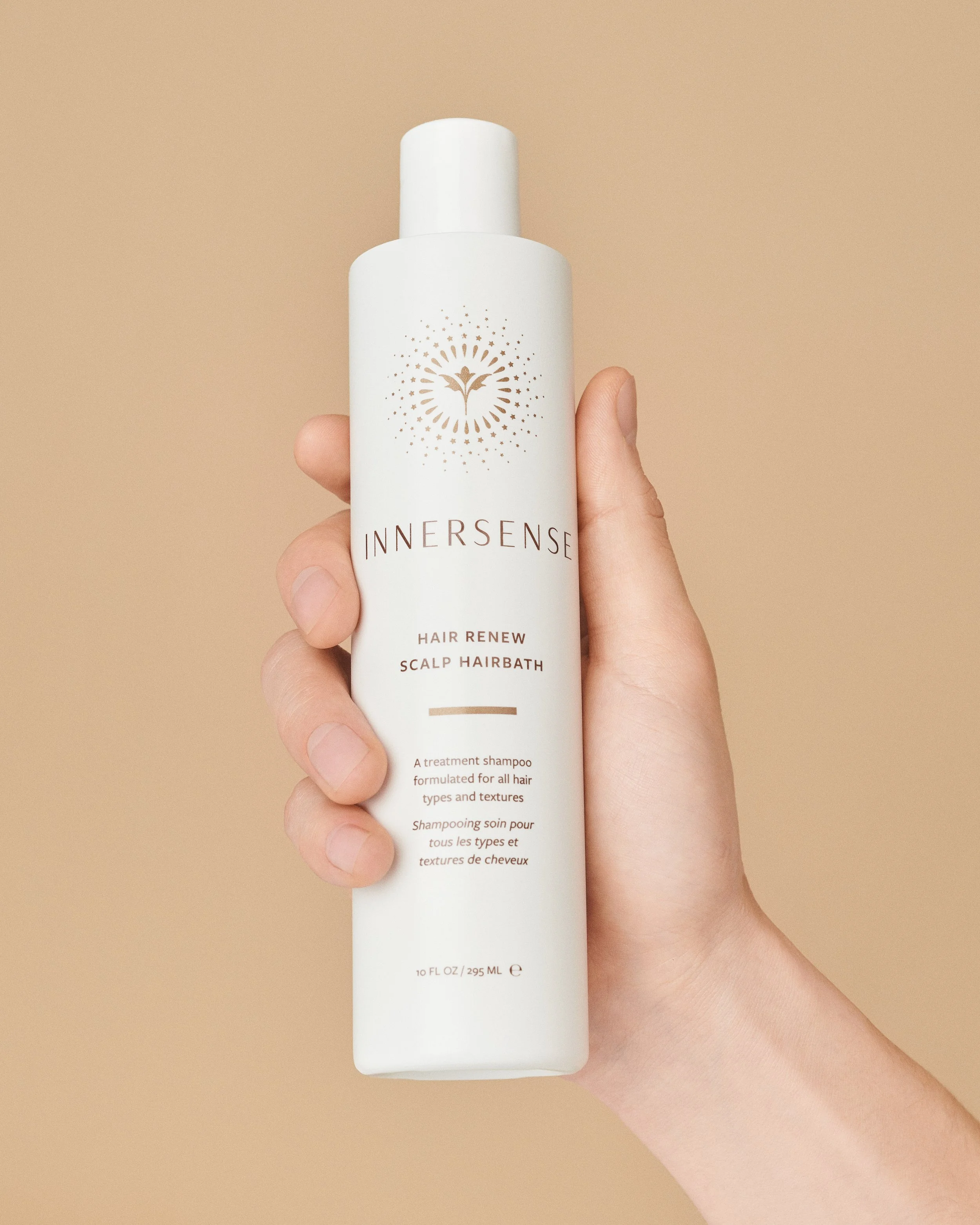 A hand holds a white bottle of Innersense Hair Renew Scalp Hairbath against a neutral beige background.