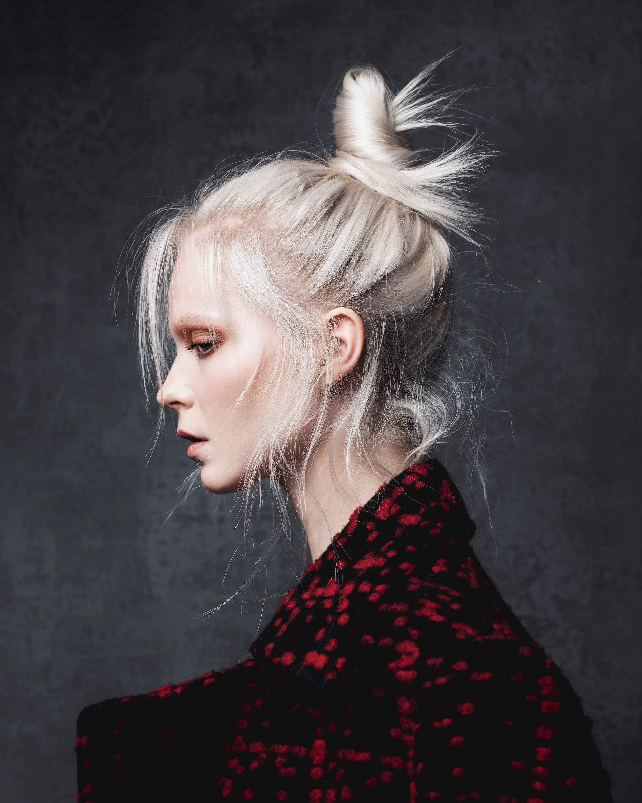 A woman with platinum blonde hair styled in a messy bun with wispy strands, wearing a black and red textured jacket, against a dark background.
