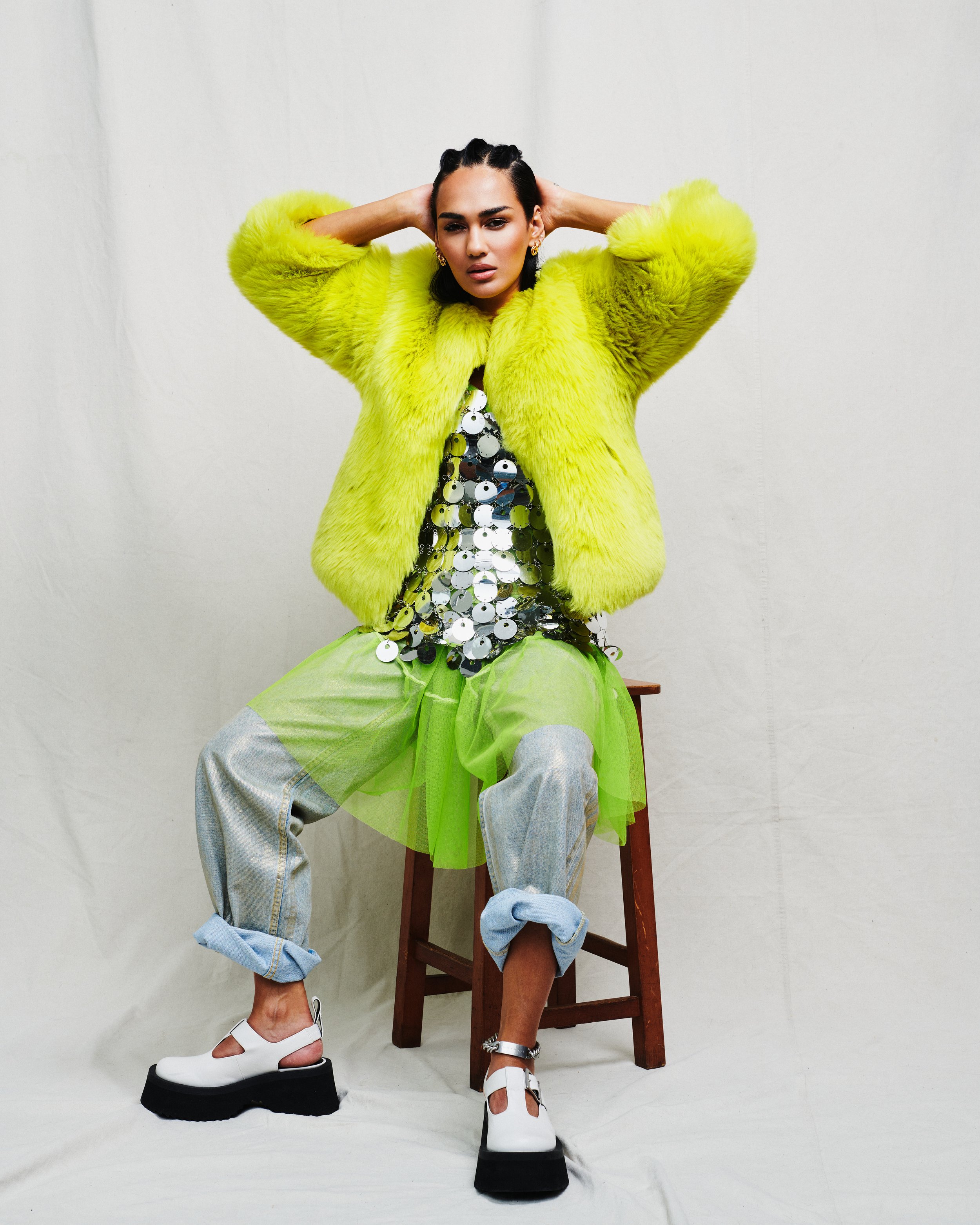 A woman in a bright yellow fur coat, shiny sequined top, oversized light blue jeans, and chunky white platform boots, sitting on a wooden chair, posing with hands behind her head against a plain background.