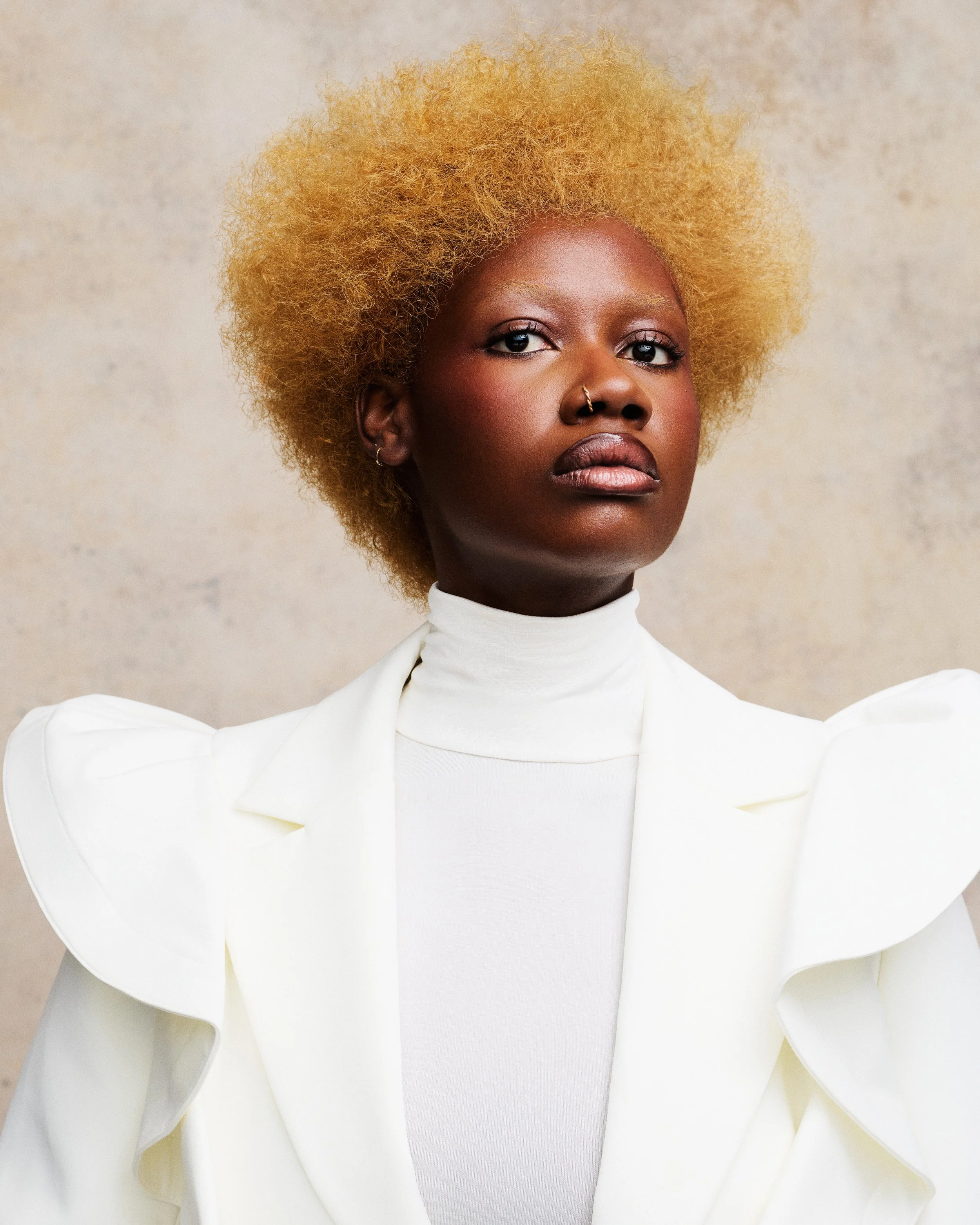 A woman with dark skin and a large, curly, blonde hairstyle posing against a plain light-colored background. She is wearing a white high-neck top with puffed sleeves and has a nose ring and small earrings.