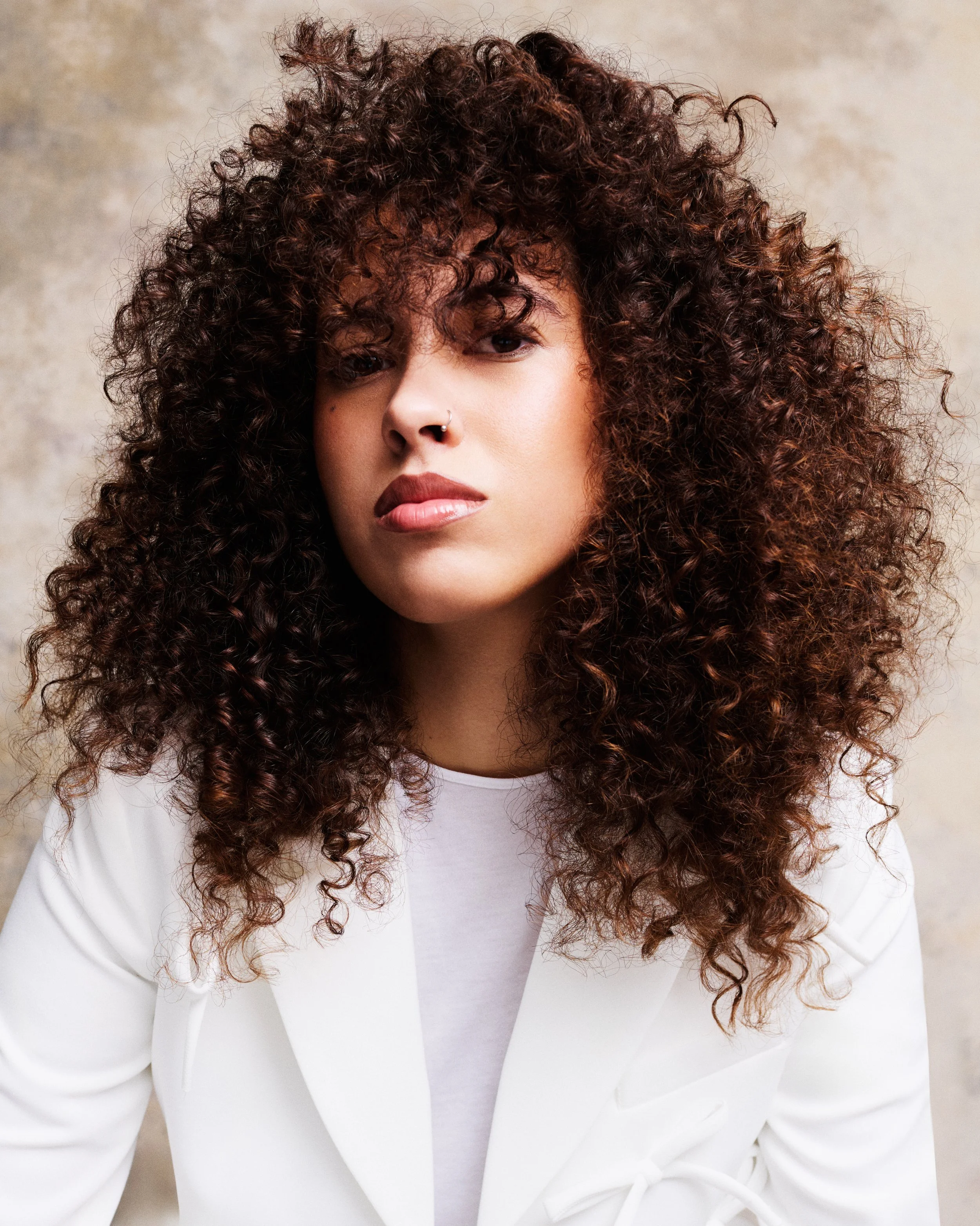 Close-up of a woman with voluminous, curly, reddish-brown hair, wearing a white blazer and a Russian-style nose ring, standing against a neutral background.