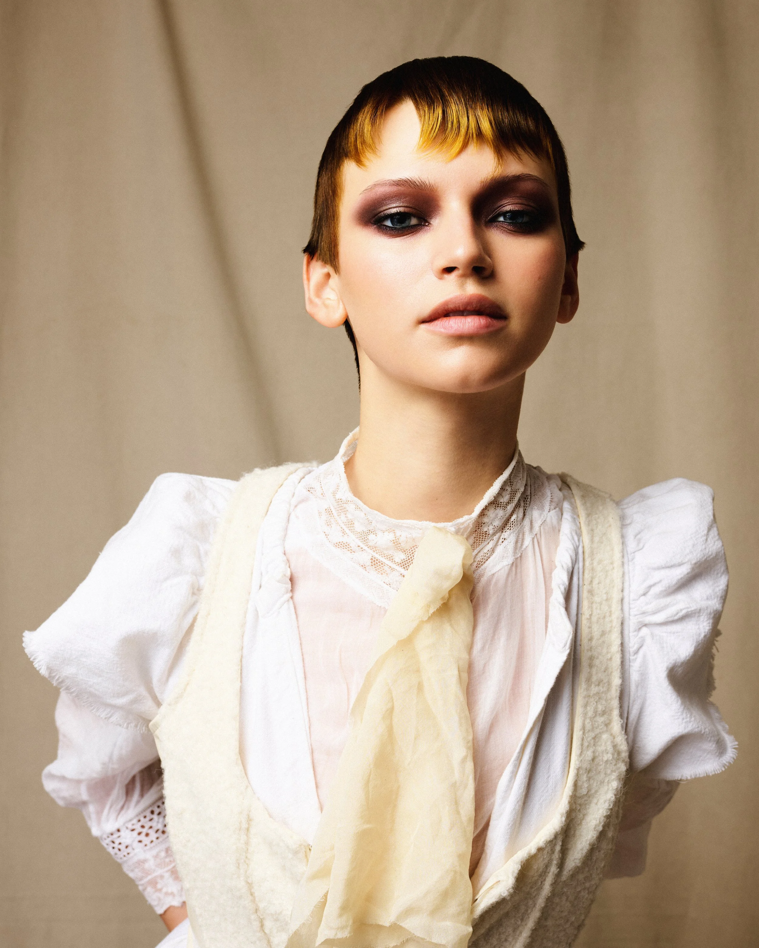 A young woman with short hair and smoky eye makeup wearing a vintage white lace blouse and cream vest, posing in front of a beige background.
