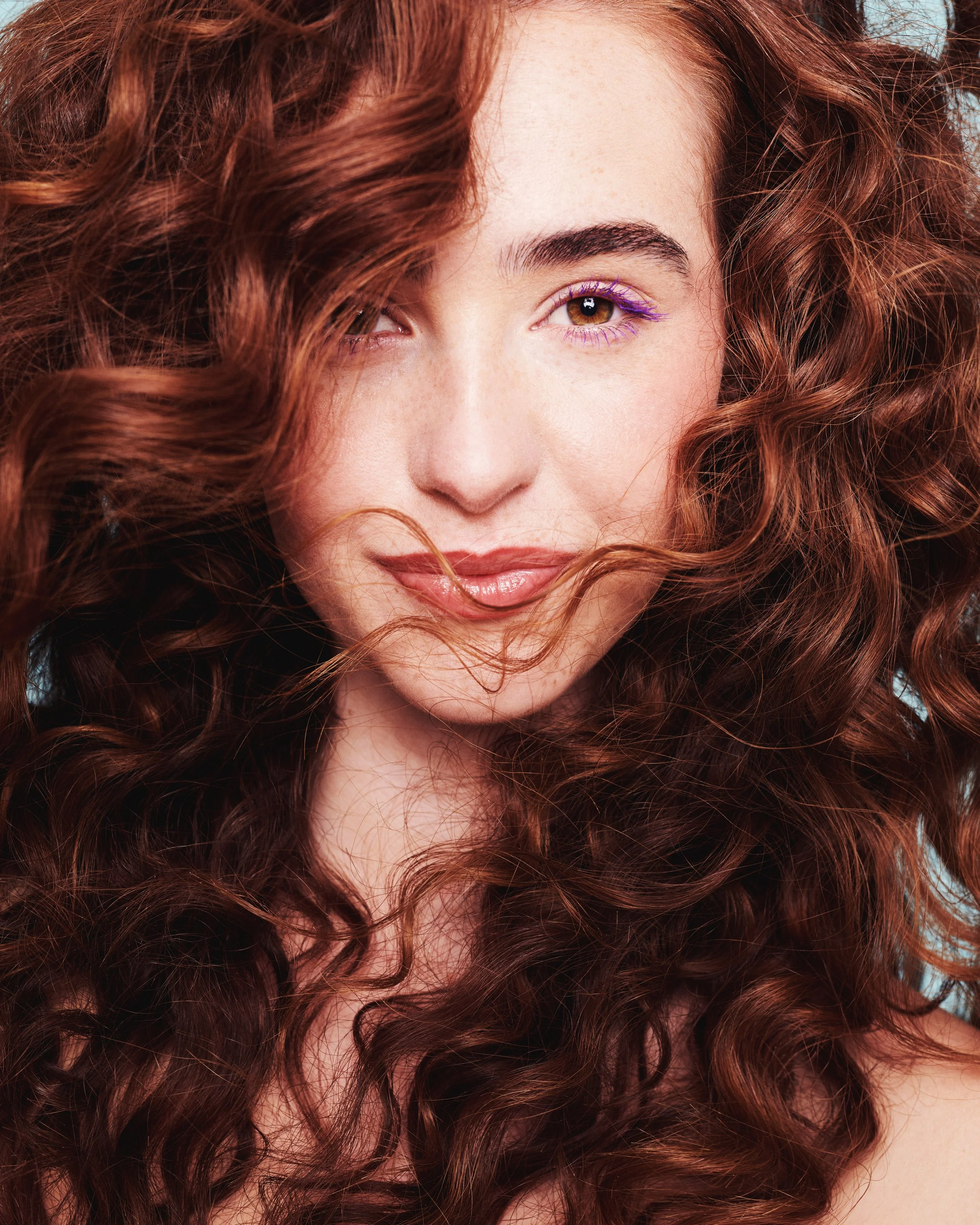 Close-up of a woman's face with curly red hair, brown eyes, and purple eyeliner.