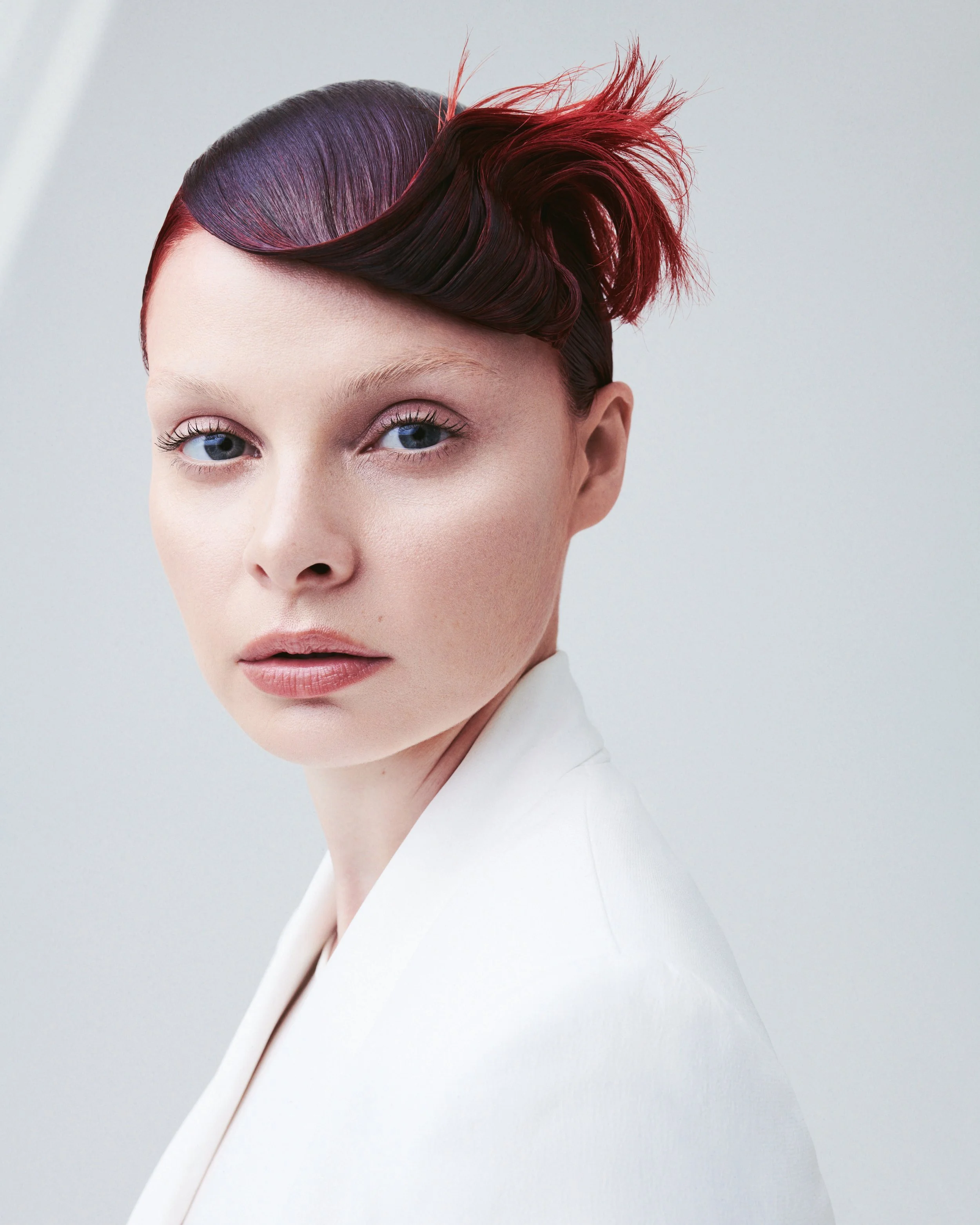 A close-up of a woman with vintage style hair, featuring a sleek, shiny dark side with purple hues and a dramatic, voluminous red updo.