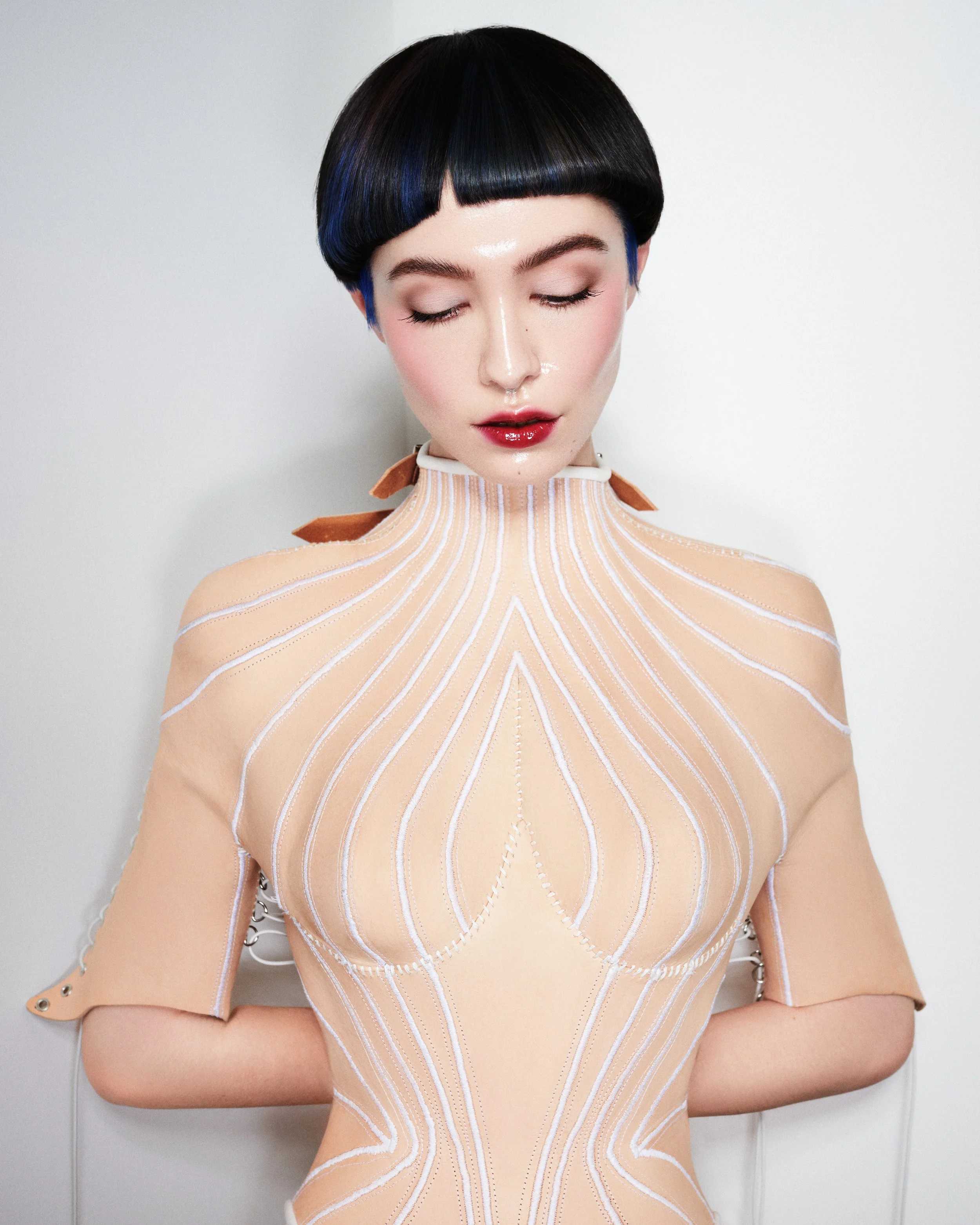 A person with short black hair styled in a bowl cut, wearing bold makeup with glossy red lipstick, and a sheer, beige high-neck top with white lines and metallic accents, standing in front of a plain white background.