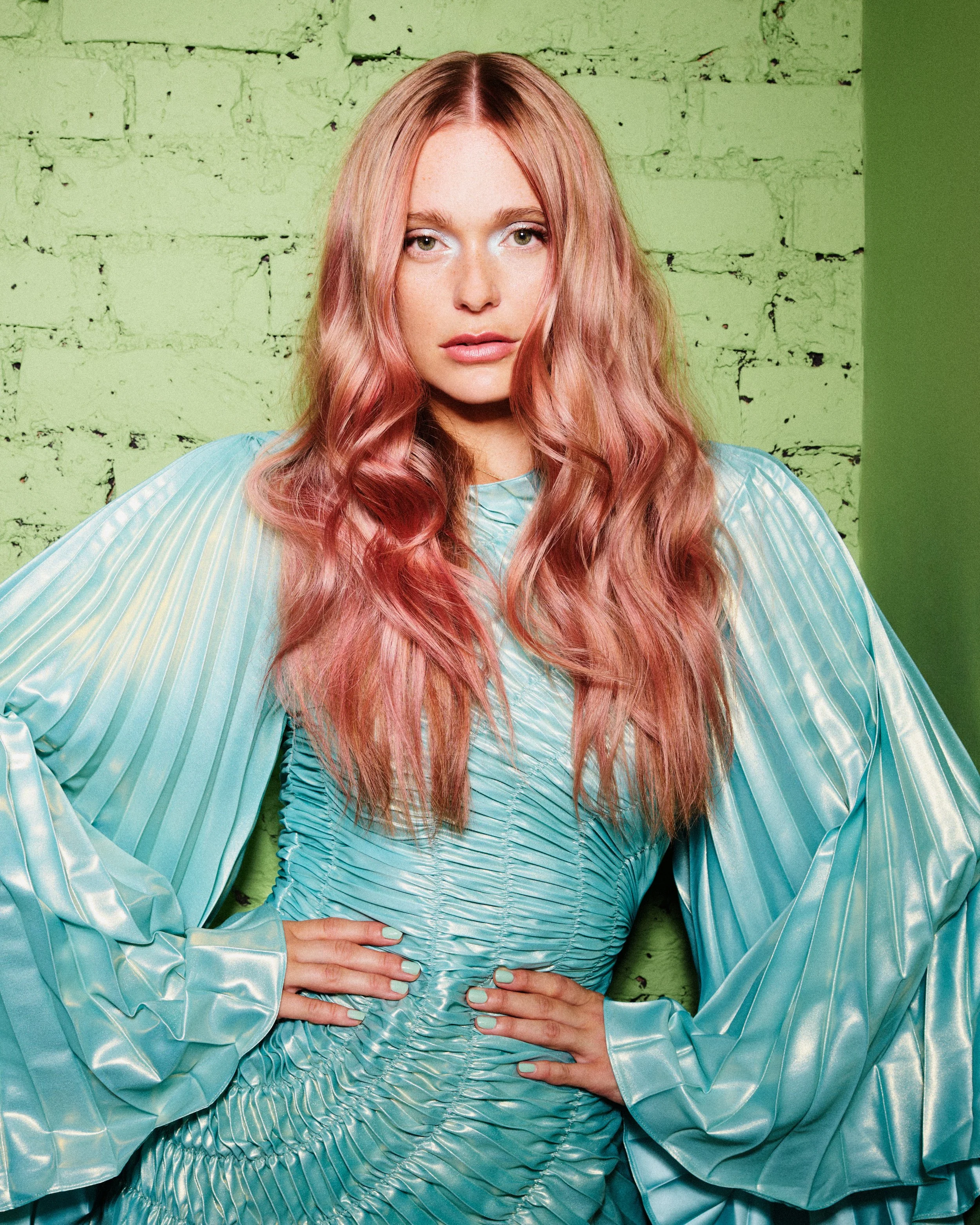 A woman with long, wavy pink hair, wearing a shiny, light blue, ruched dress and matching gloves, poses with her hands on her hips against a green brick wall background.