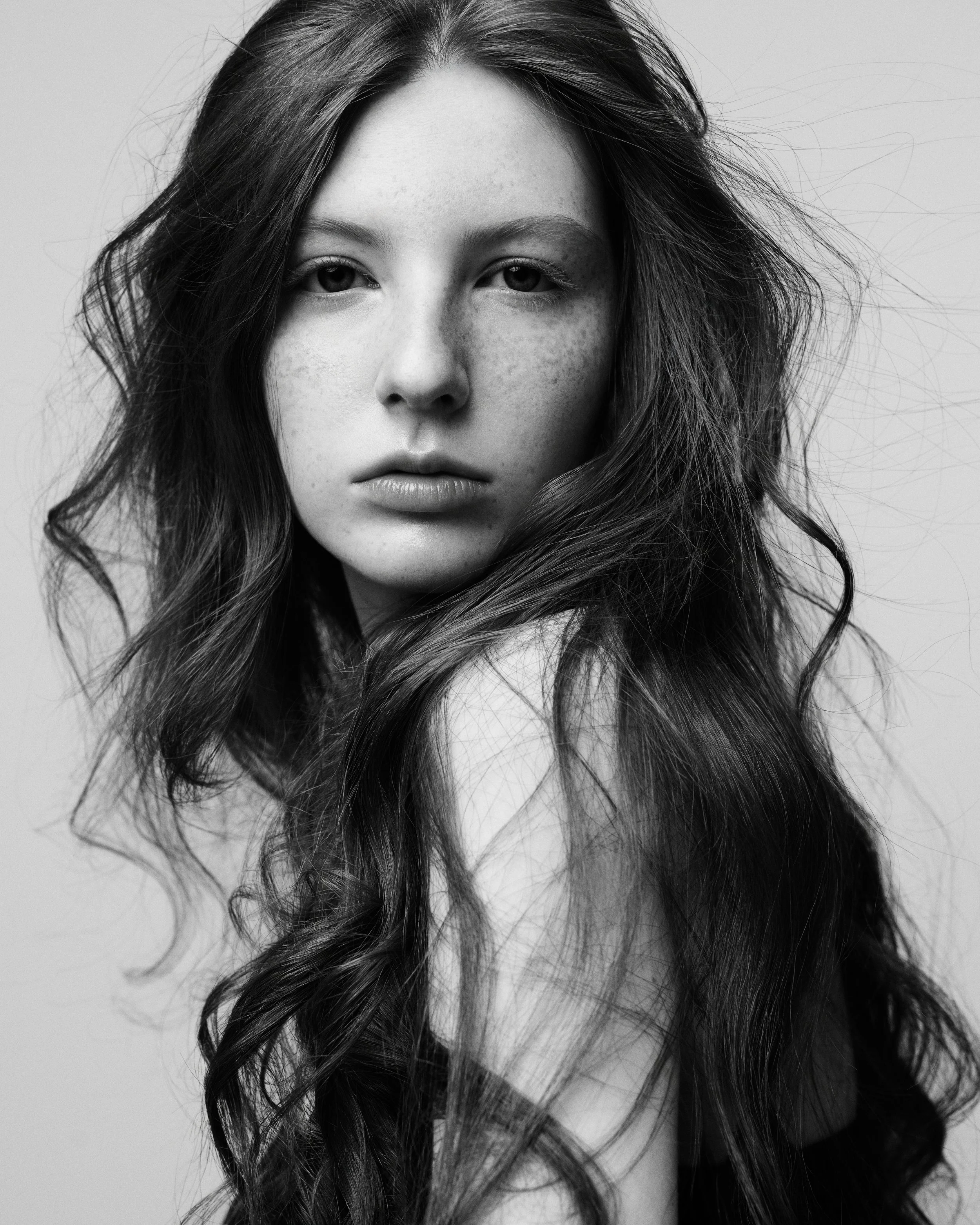 Close-up black-and-white portrait of a young woman with long, wavy hair and a neutral expression, looking directly at the camera.