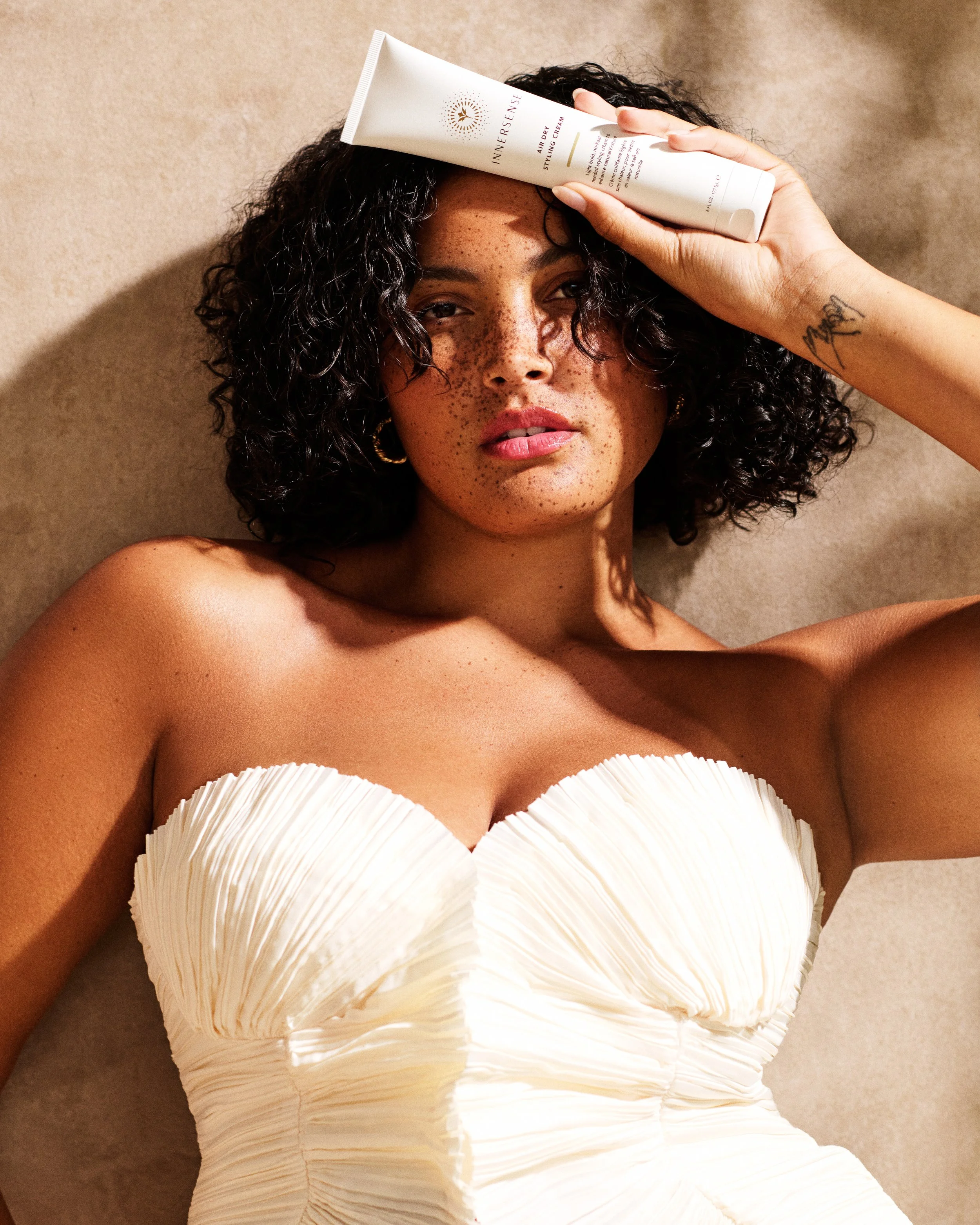 A woman with curly dark hair and freckles lying on a beige surface, holding a white tube of Innersense Air Dry Styling Cream on her forehead, wearing a textured white strapless dress.