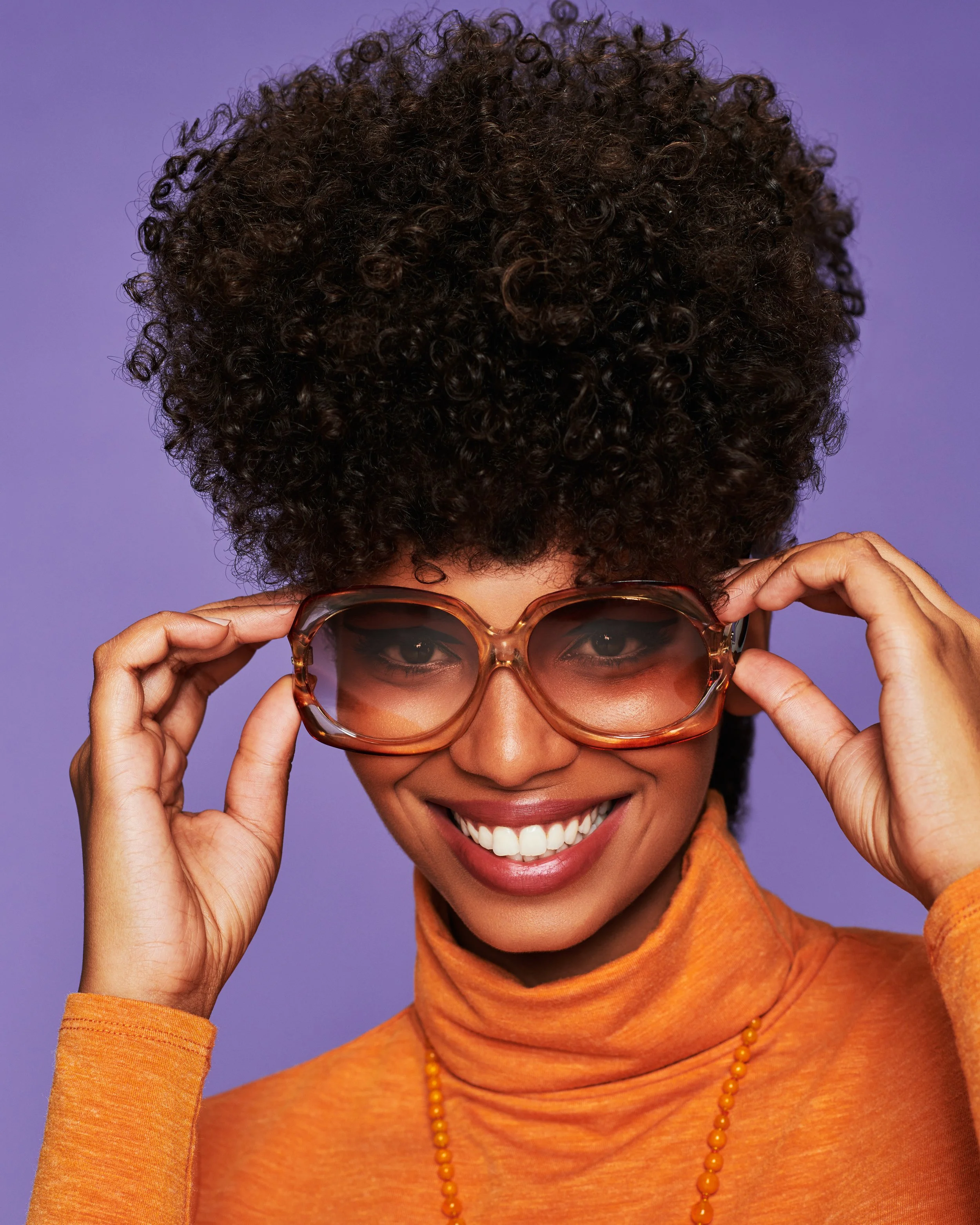 A woman with dark, curly hair smiling, wearing orange sunglasses, an orange turtleneck, and an orange beaded necklace, adjusting her glasses with her hands, against a purple background.