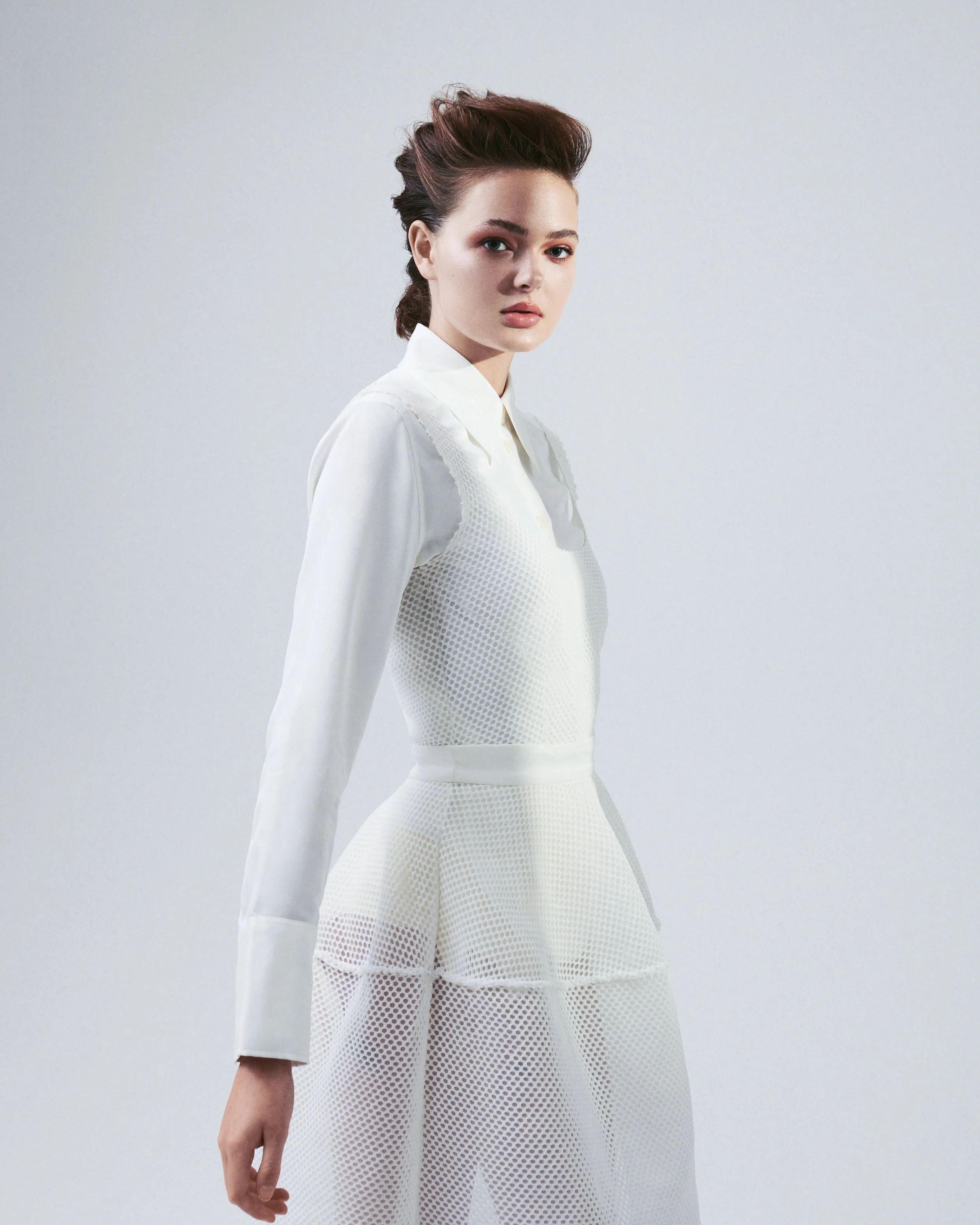 A woman in a white outfit with long sleeves and a high-waisted skirt, standing against a plain light gray background, with brown hair styled in a voluminous updo.