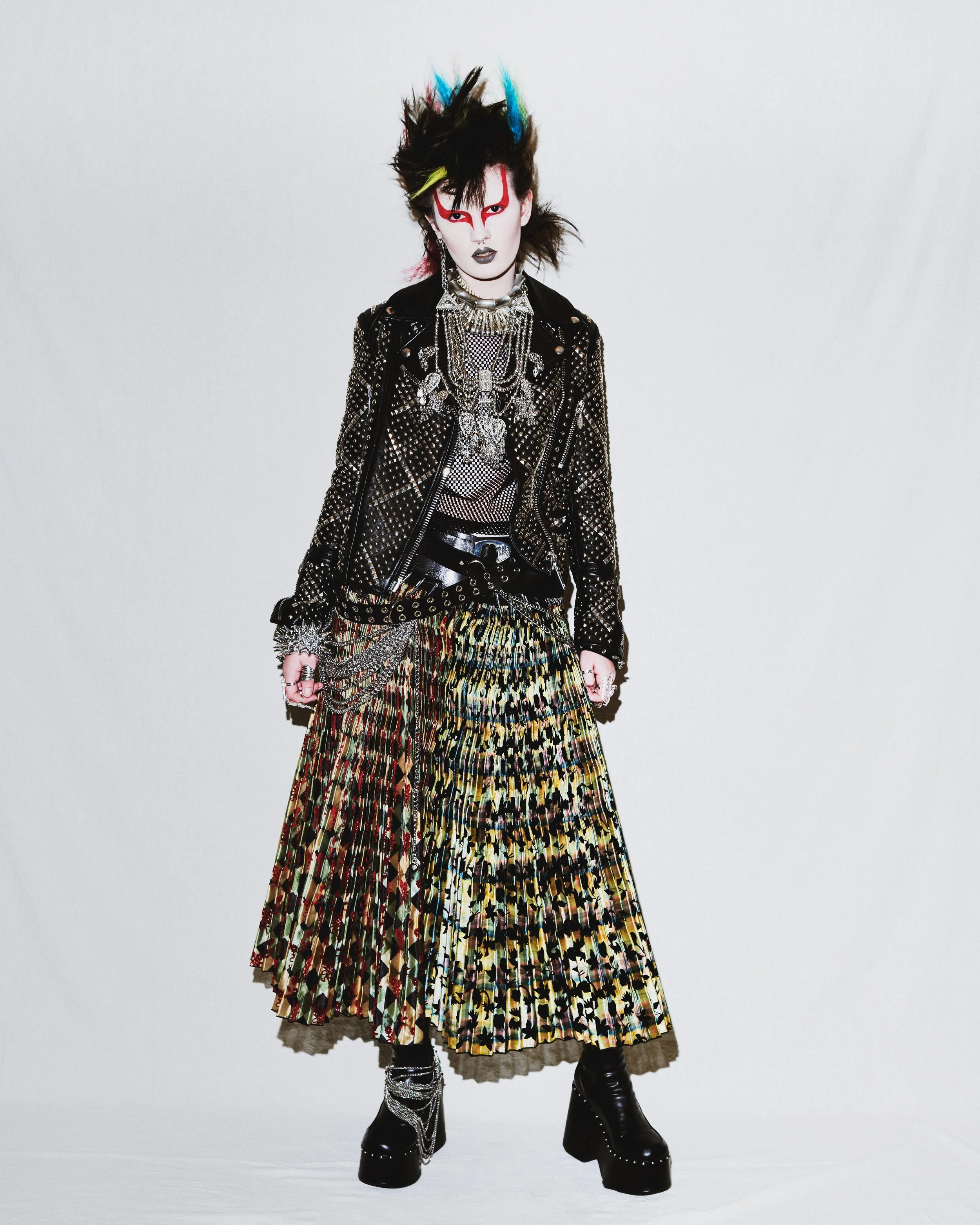 Person with spiked multicolored hair, white face makeup with red designs, black lipstick, wearing a black studded leather jacket, layered silver necklaces, a mesh top, a colorful pleated skirt, and platform black boots with chains and studs.