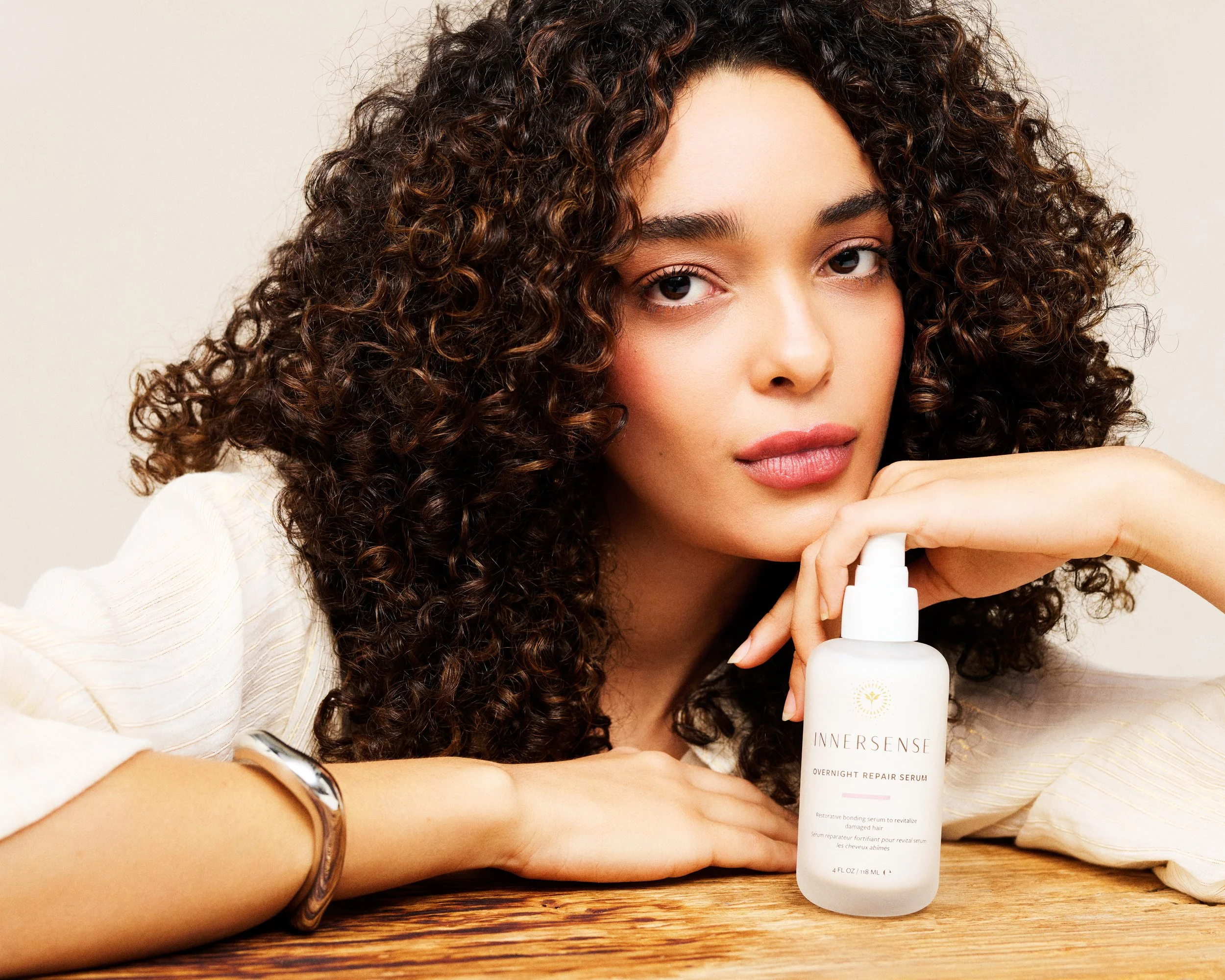 A woman with curly brown hair holding a white bottle of Innersense Overnight Repair Serum, resting her chin on her hand, with a wooden table in front of her.