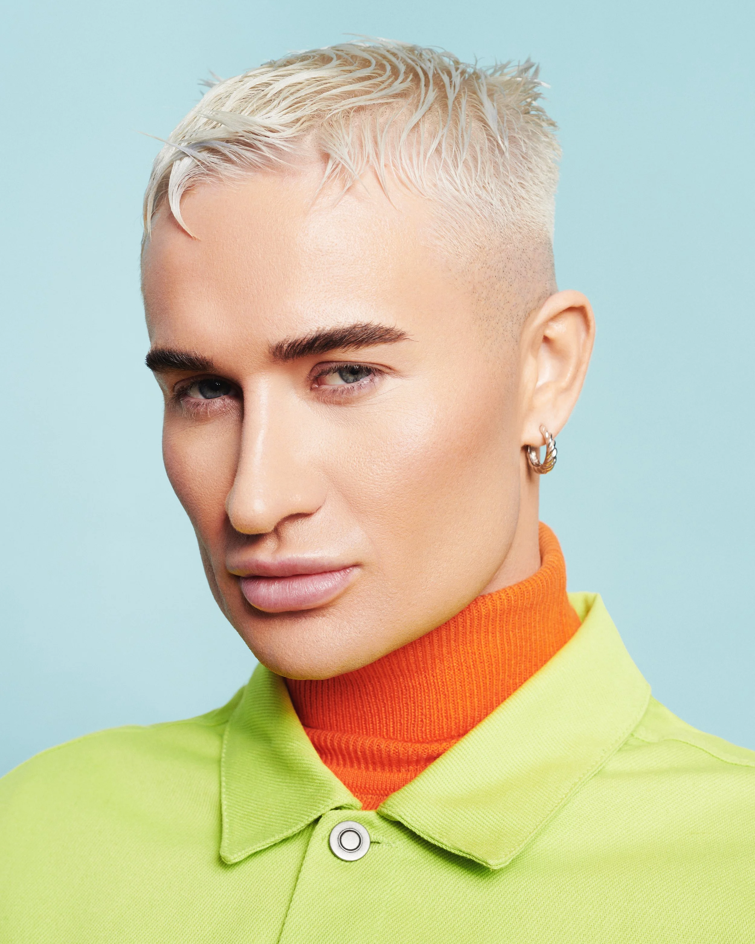 Close-up of a young person with short platinum blonde hair styled upward, wearing a lime green jacket over an orange turtleneck, against a light blue background.