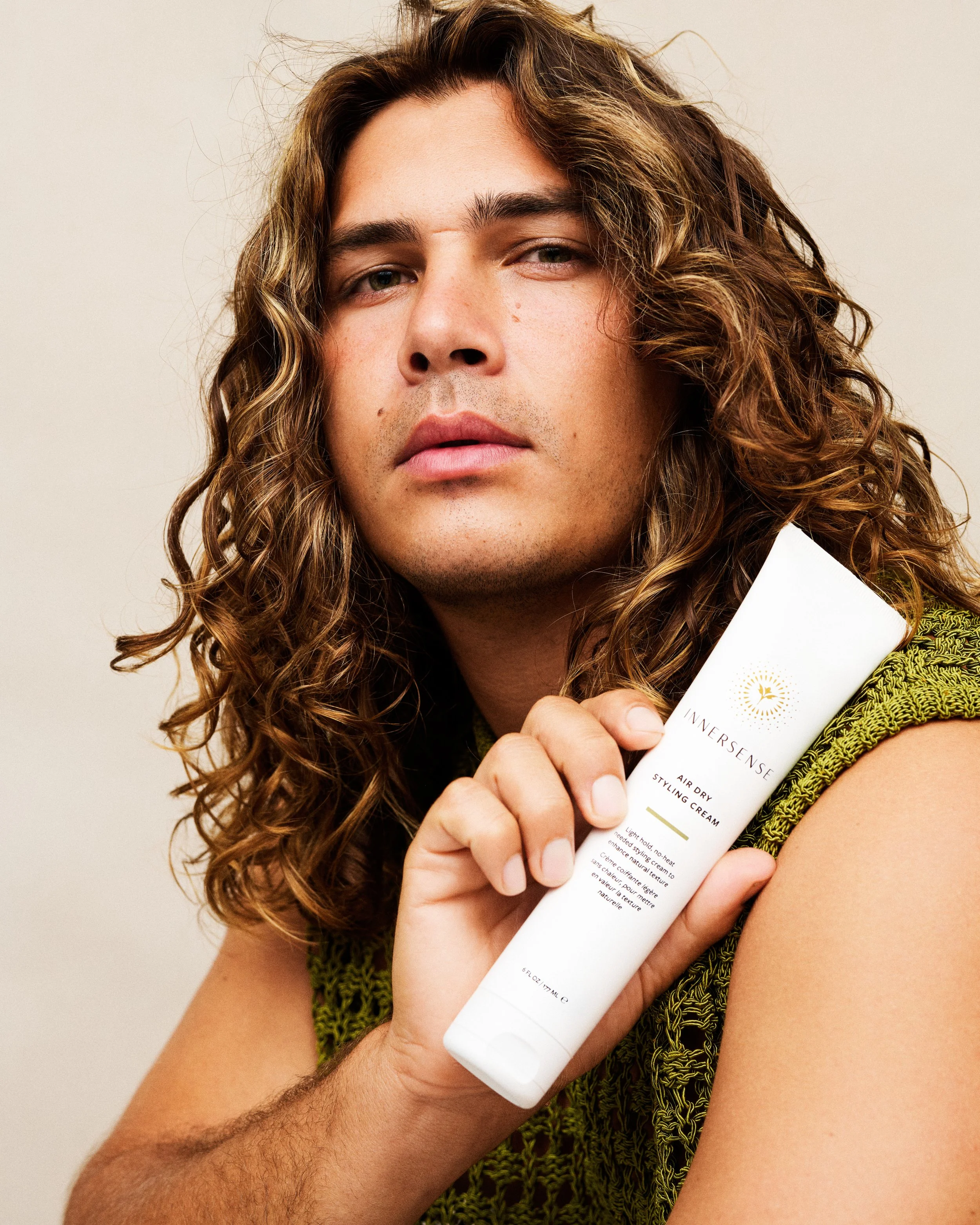 A man with curly hair holding a white tube of skincare or cosmetic product close to his face, wearing a green textured top, against a neutral background.