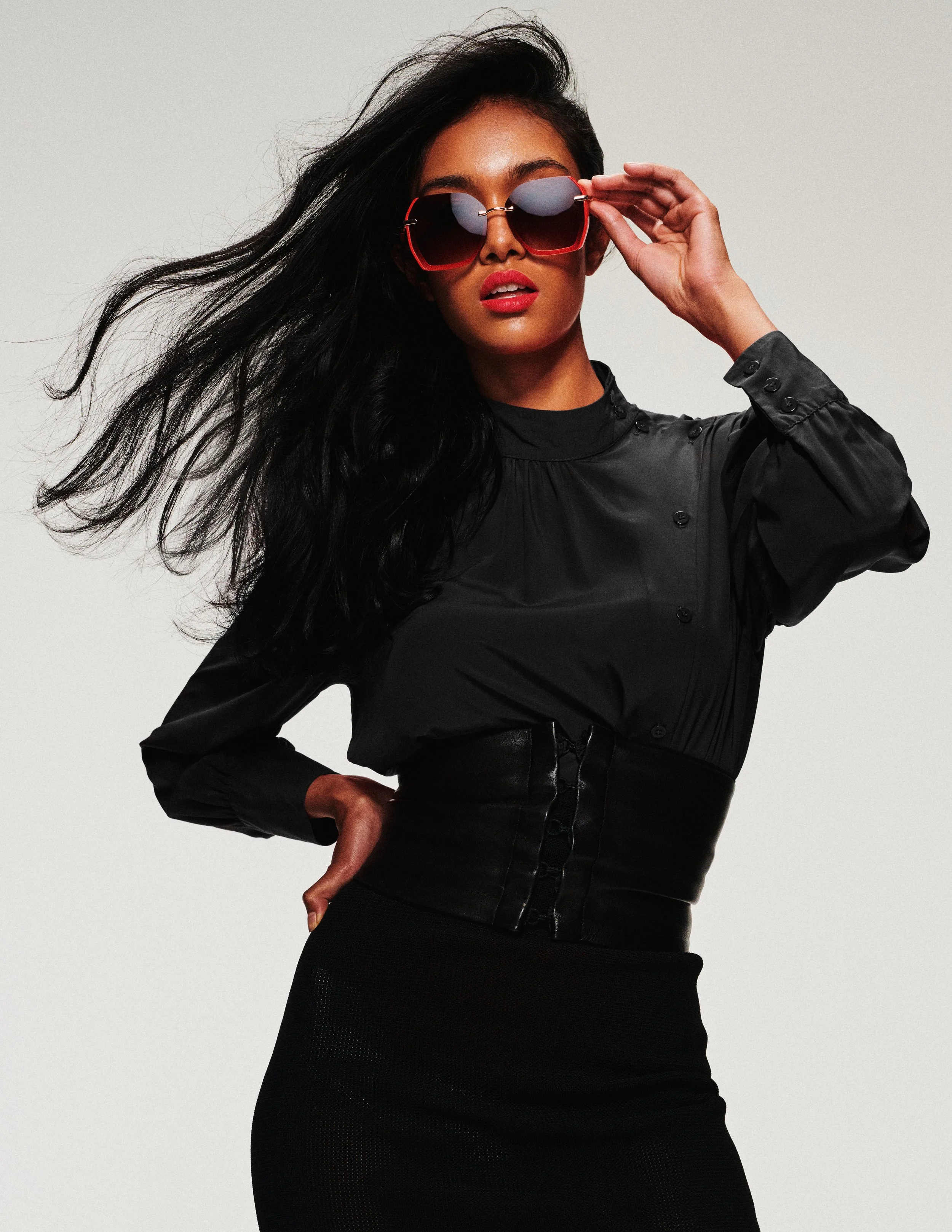 A woman with long black hair wearing red sunglasses, a black button-up shirt, and a black skirt, posing with one hand on her hip against a plain white background.