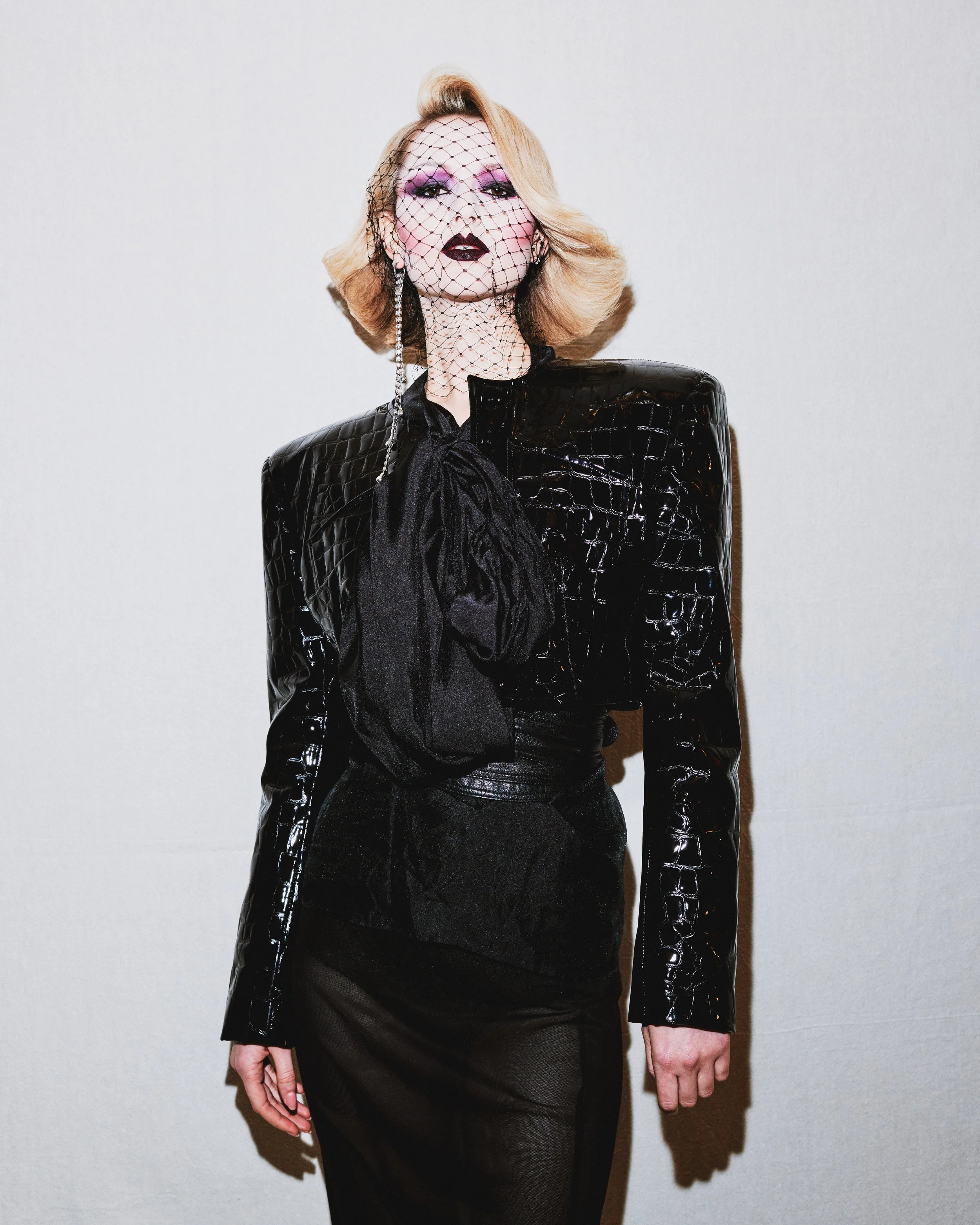 A woman with blonde hair styled in a vintage wave, wearing dark lipstick and purple eye makeup, dressed in a black leather-like textured jacket with a black satin bow tie and long black gloves, standing against a plain white wall.