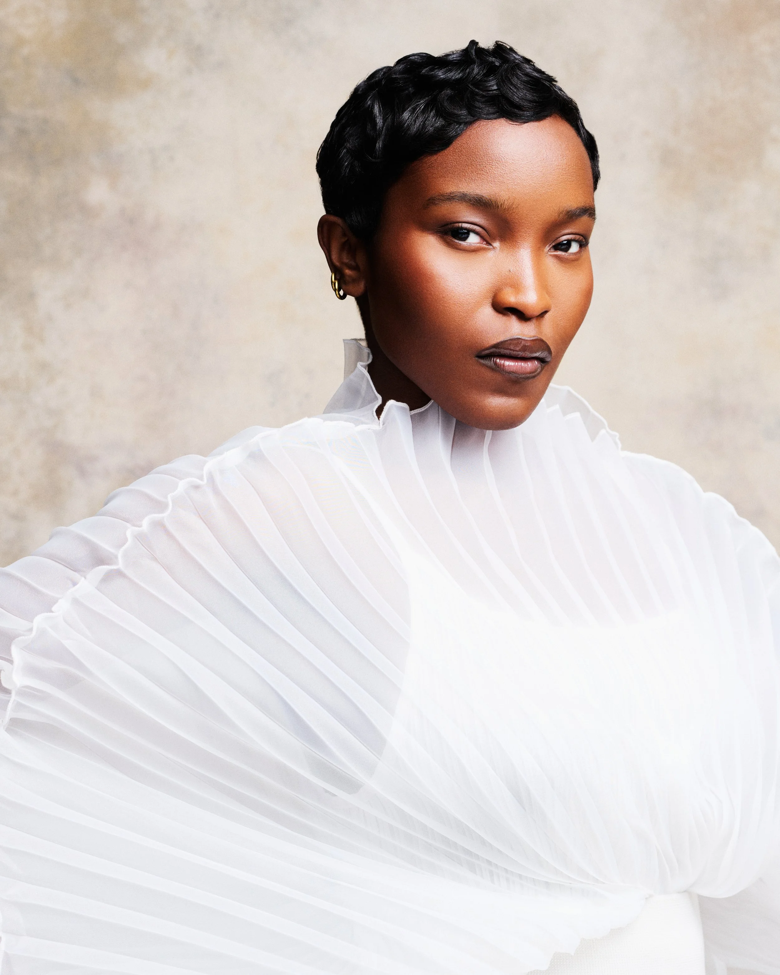 Close-up portrait of a woman with short black hair, wearing a white ruffled blouse and gold earrings, looking confidently at the camera against a neutral textured background.