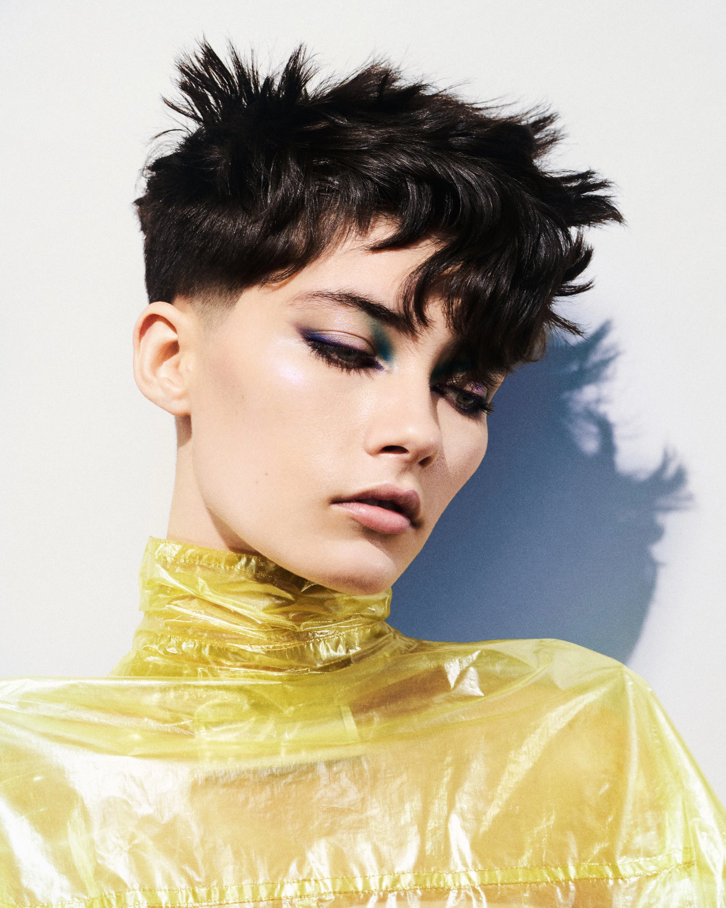 A close-up of a person's short, dark, and layered hairstyle with a drooping slightly tousled top. The individual has fair skin, subtle makeup with purple and green eye shadow, and a glossy finish on their skin. They are wearing a yellow, shiny, crink