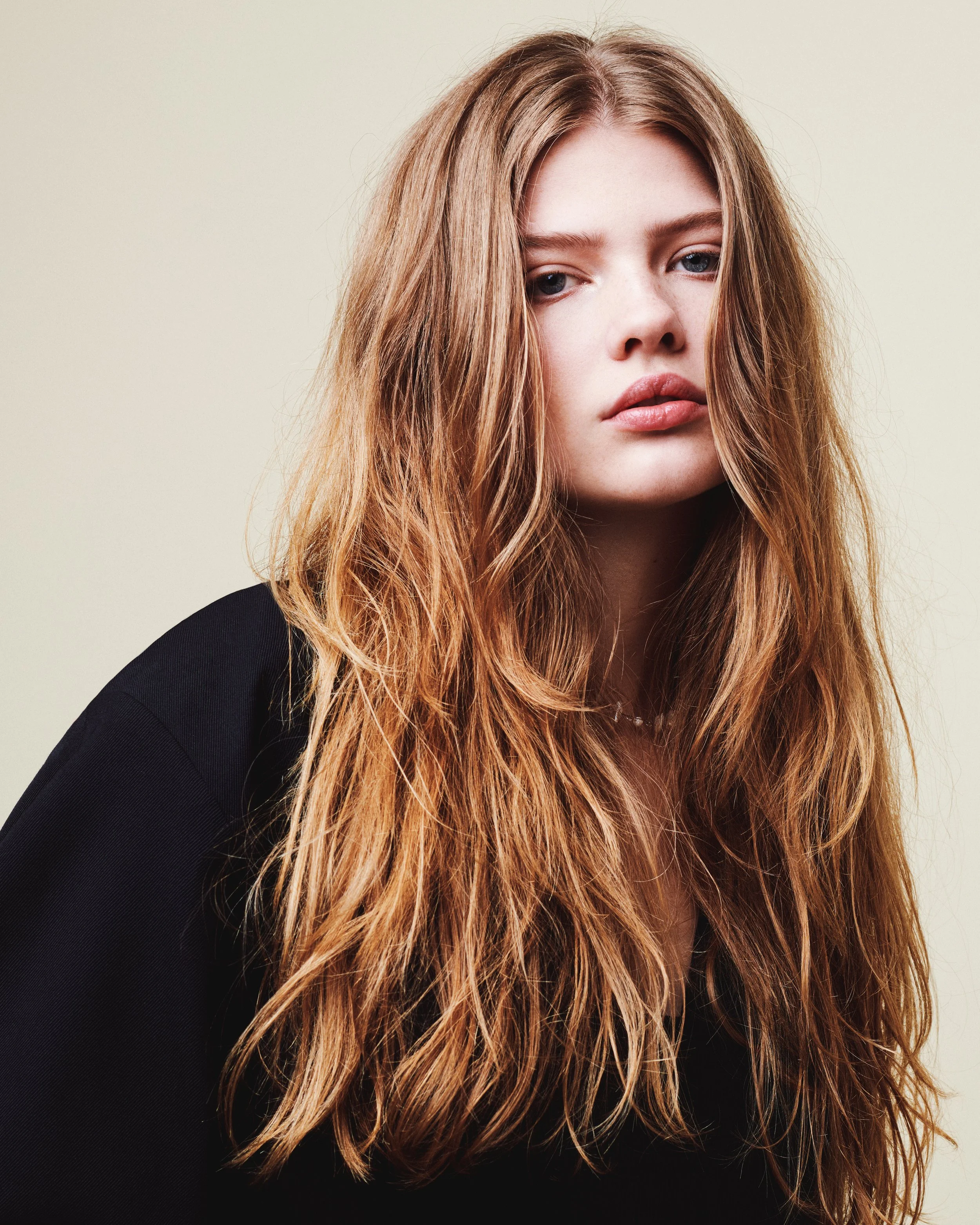 Close-up of a young woman with long, wavy, blonde hair and blue eyes, wearing a black top, against a plain light background.