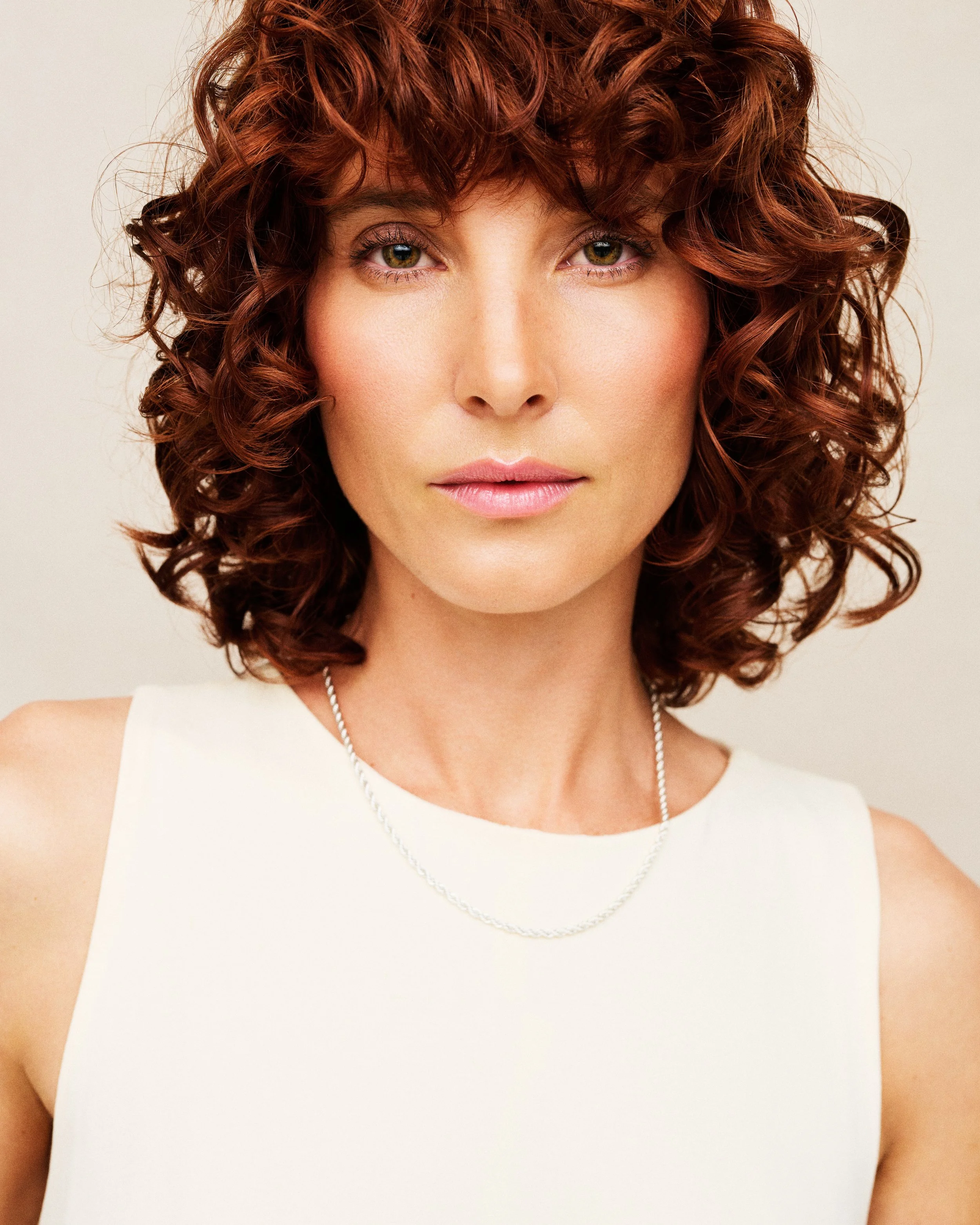 A woman with curly red hair and light makeup wearing a white sleeveless top and a silver necklace, posing in front of a neutral background.