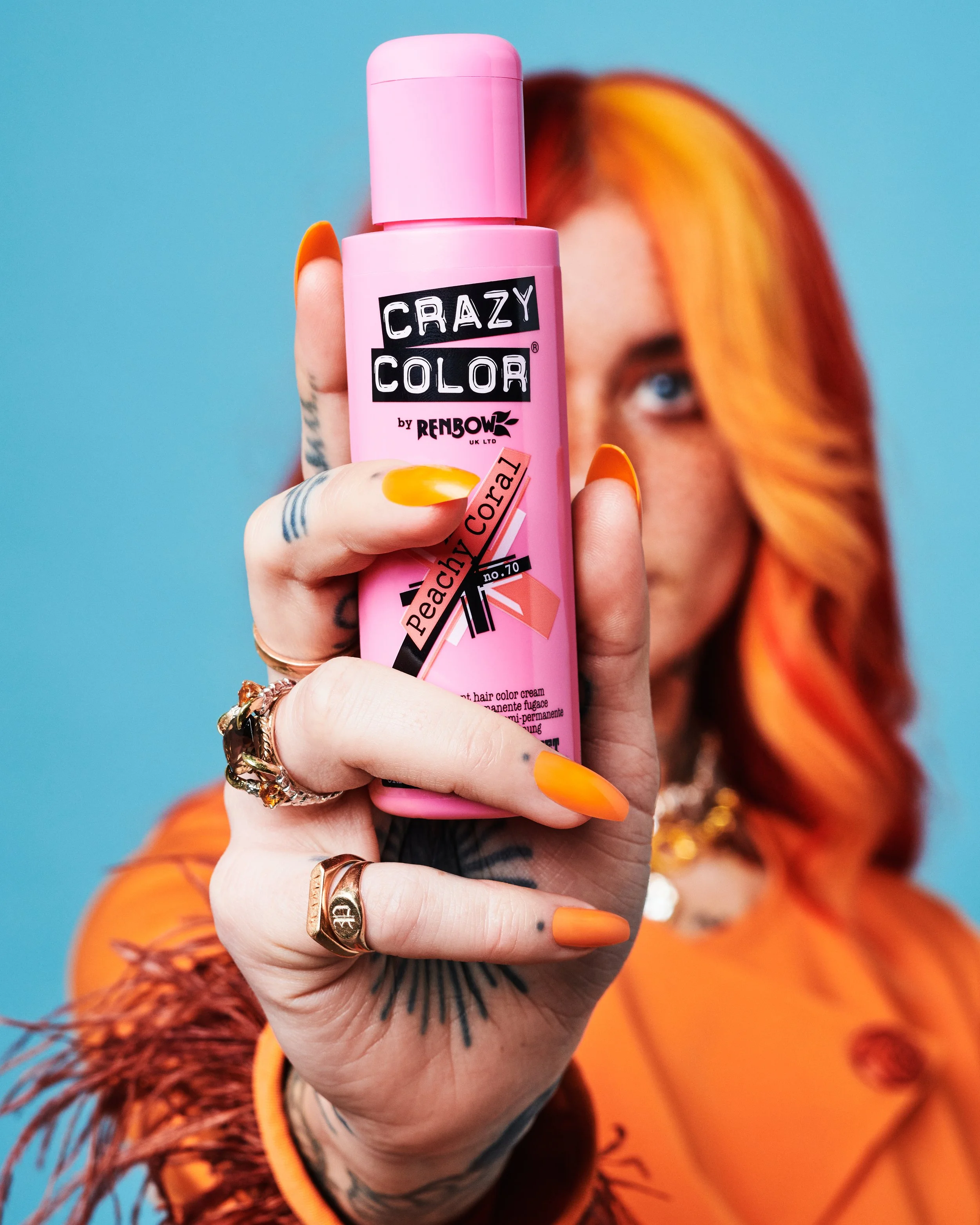 A person with orange hair and orange clothing holding a pink bottle of Crazy Color hair dye. The person's hand has multiple rings and tattoos, and the background is blue.