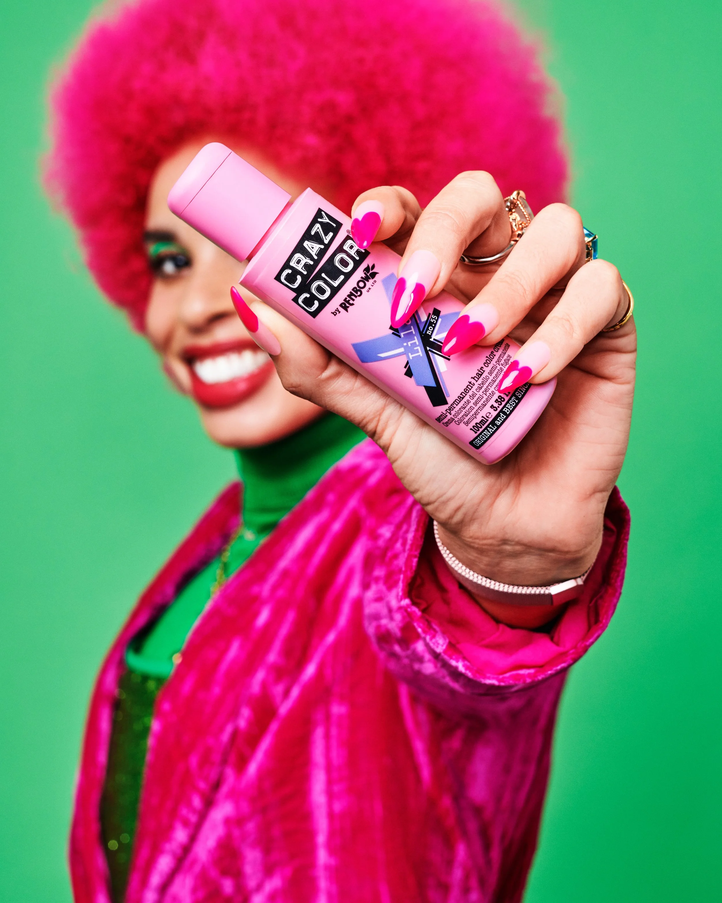 Close-up of a woman with bright pink hair, wearing pink lipstick and holding a pink bottle of hair color spray, with a colorful background.