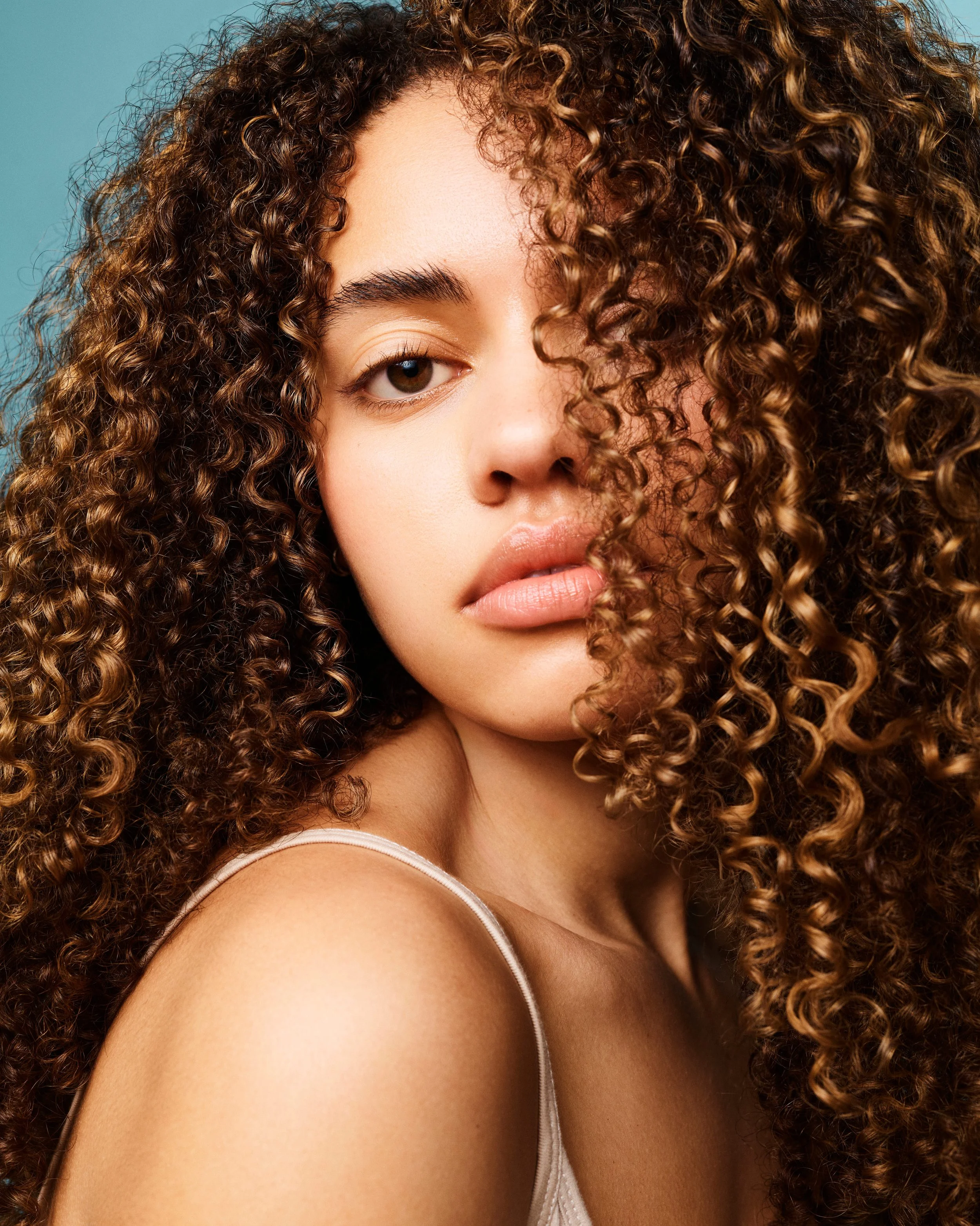 Close-up of a woman with curly brown hair, partially covering one eye, and wearing a white strap top.