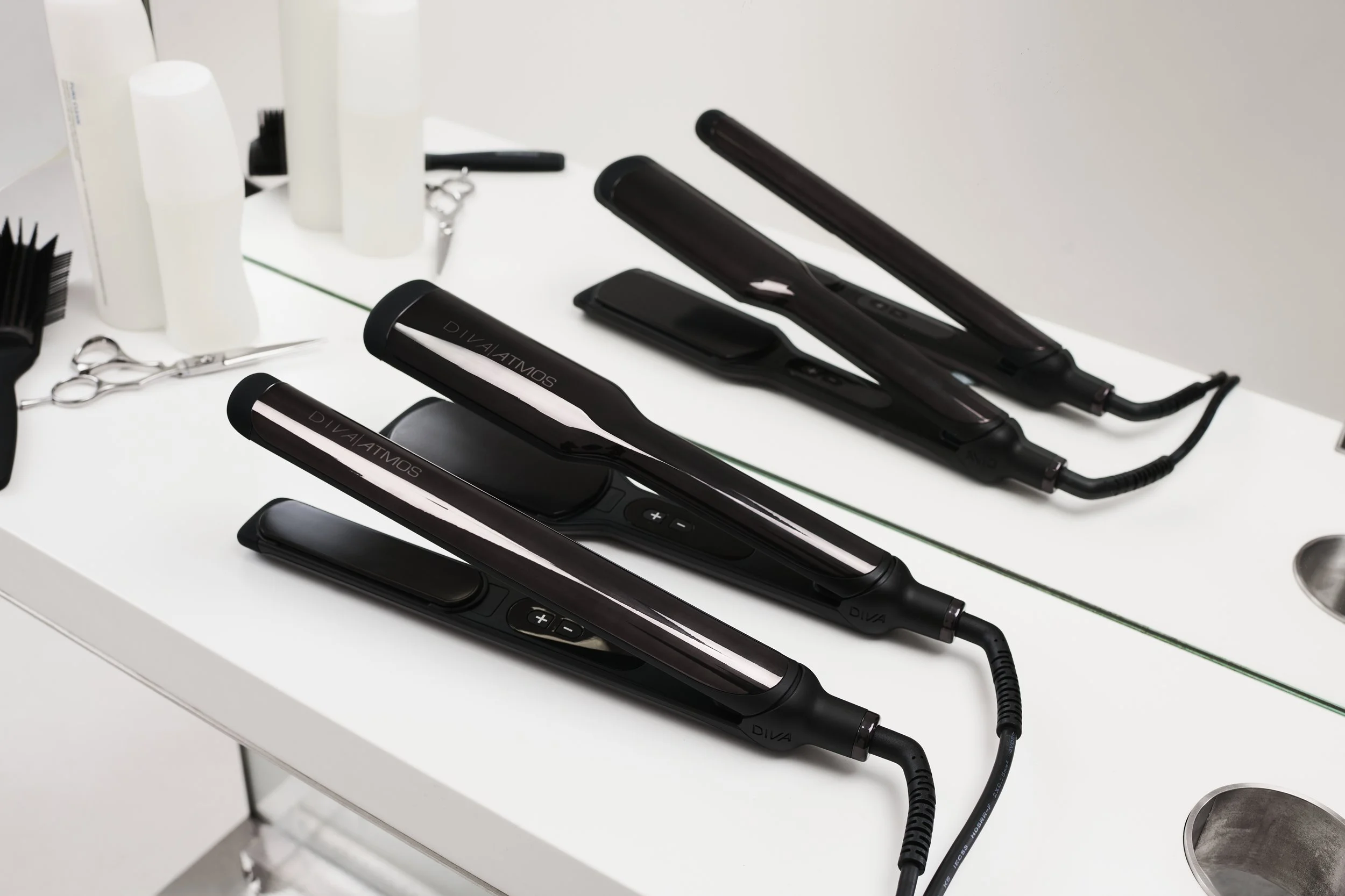 Three black flat irons with digital displays and a coiled cord are lined up on a white countertop in a hair salon, with hair scissors, white bottles, and black hairbrushes nearby.