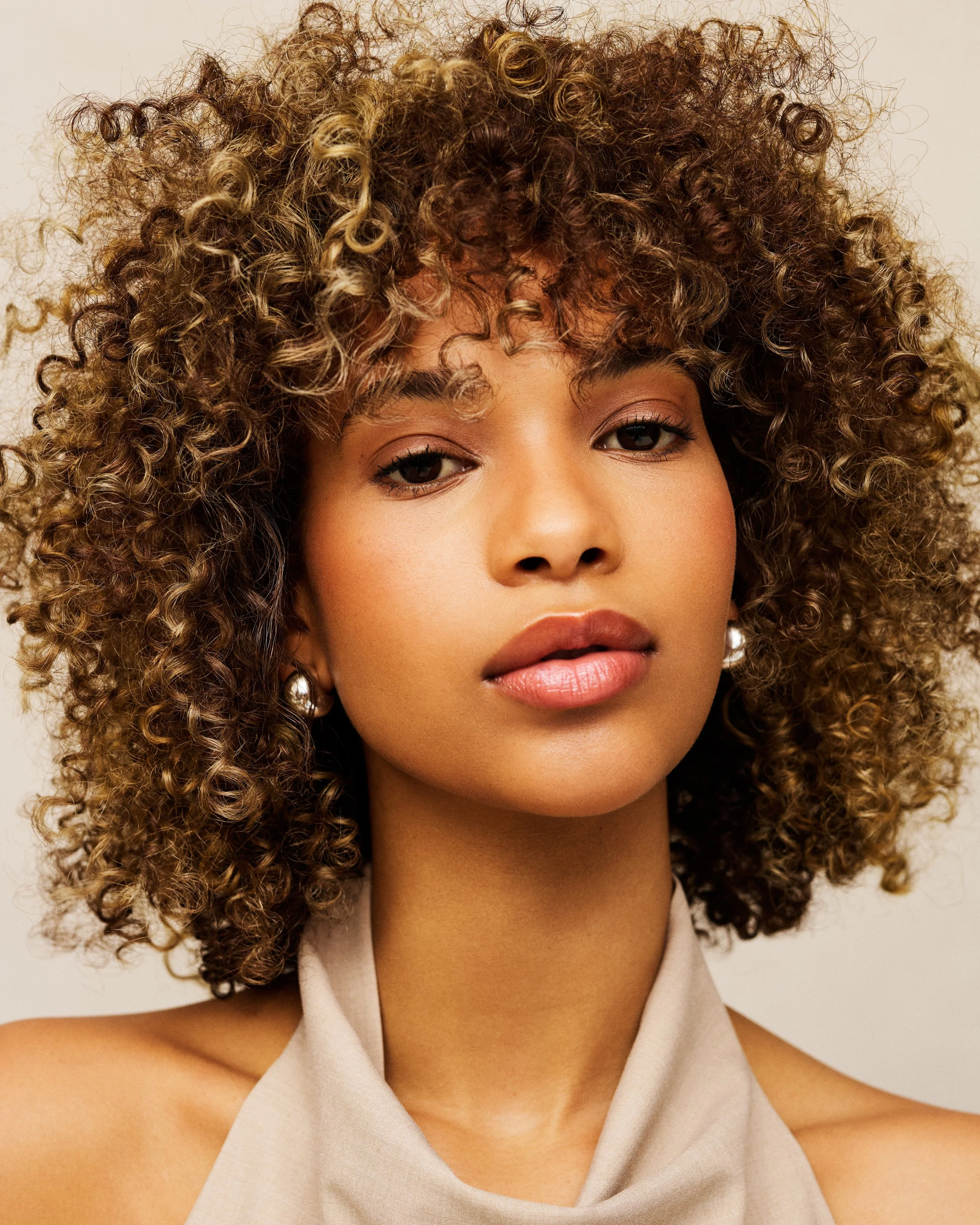 Close-up of a woman's face with curly, blonde hair and a beige sleeveless top.