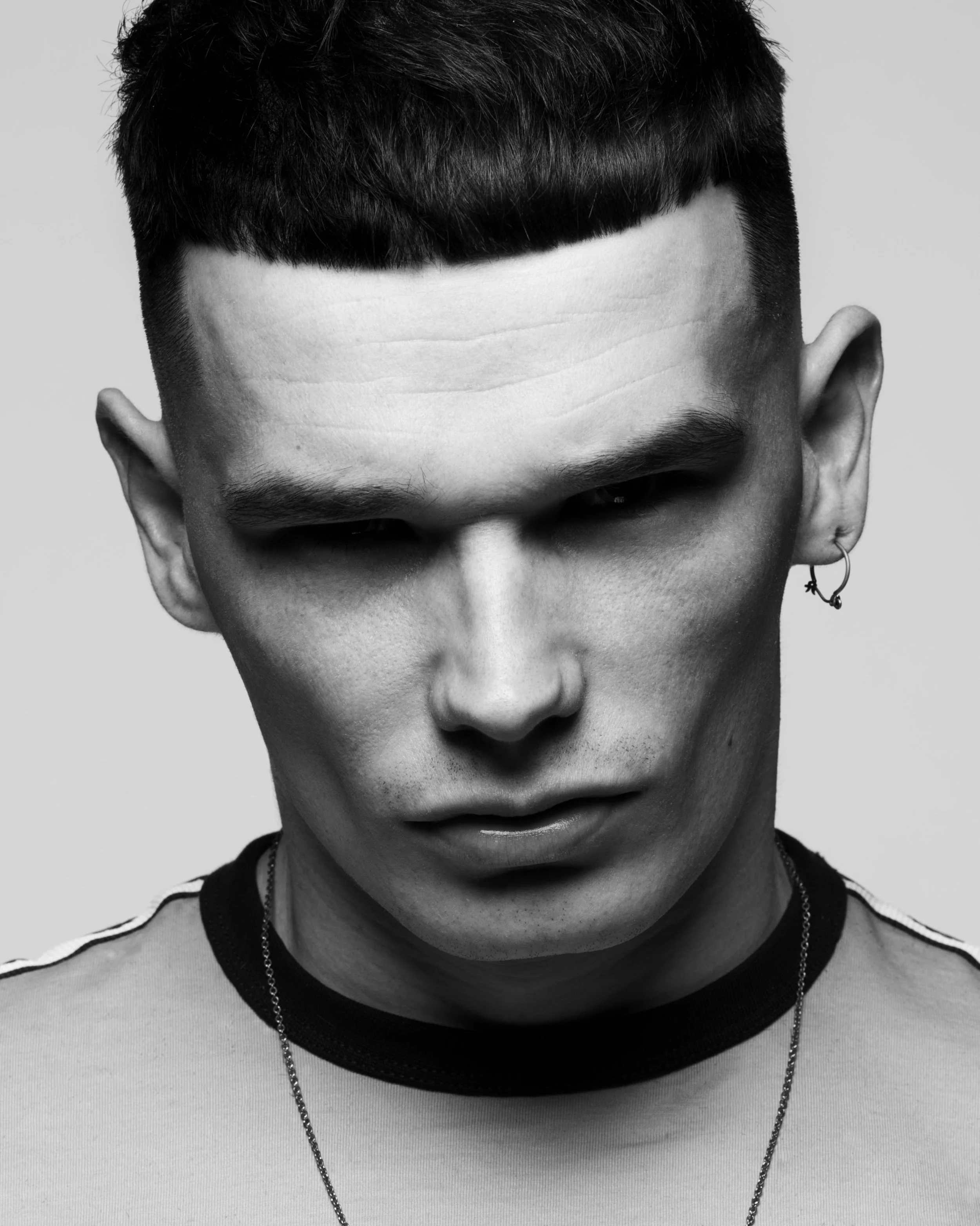 Close-up black and white photo of a young person's face, focusing on the forehead, eyes, and nose with short dark hair, an earring, and layered necklaces.