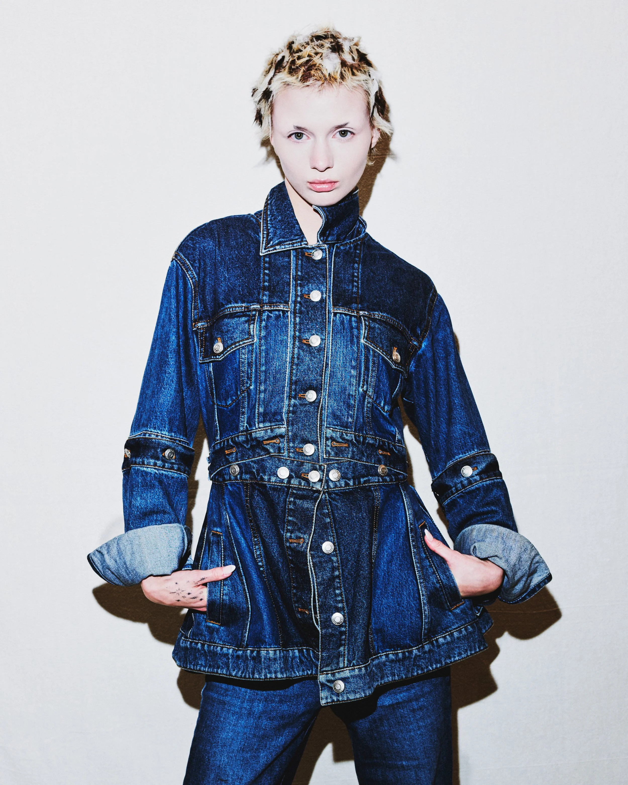 A young person with short, blond, curly hair wearing a denim jacket and jeans, with crossed arms, standing against a plain white background.