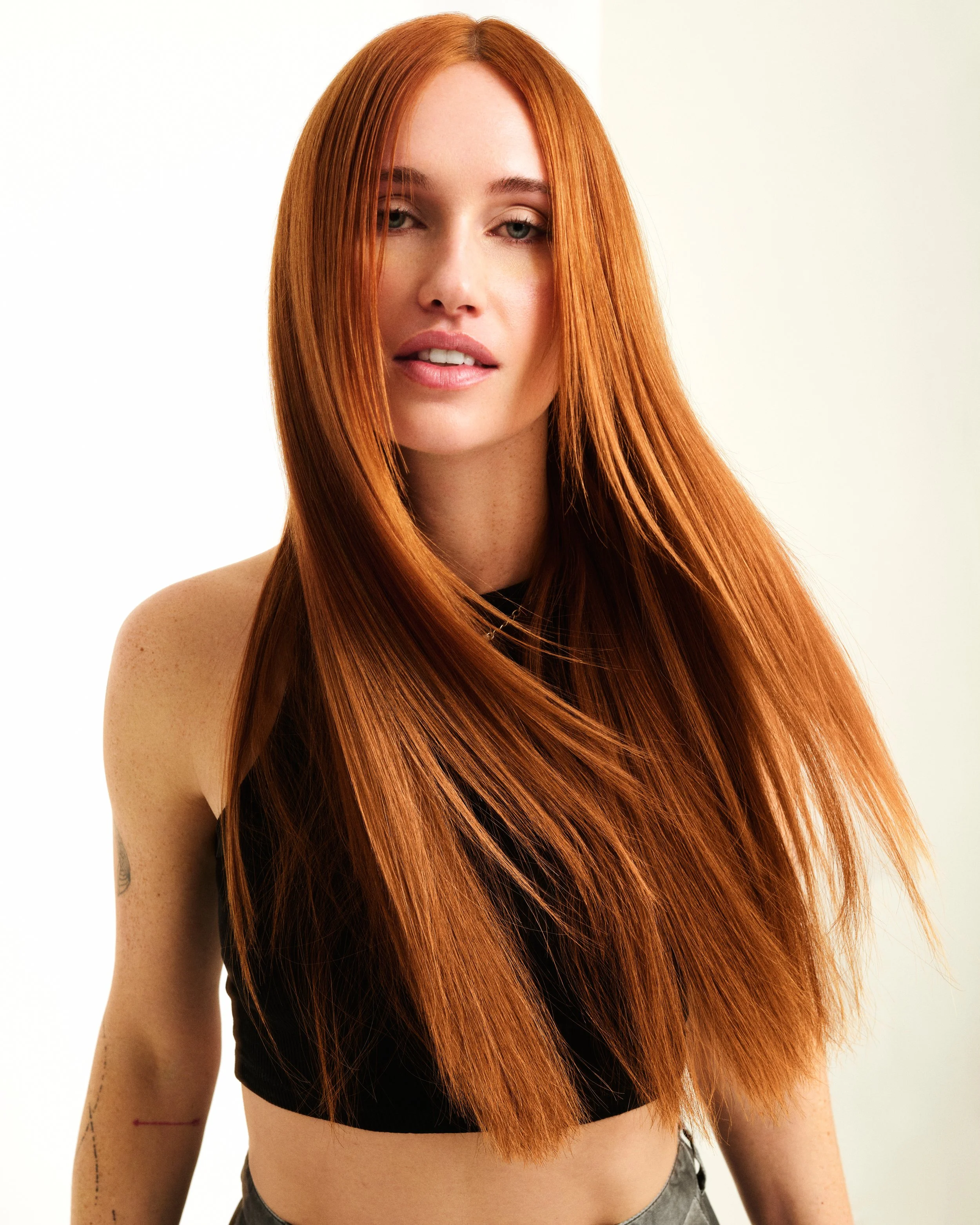 A young woman with long, straight red hair, light skin, and blue eyes looking at the camera with a relaxed expression. She is wearing a black top, with her right shoulder visible, and a tattoo on her arm. The background is plain white.
