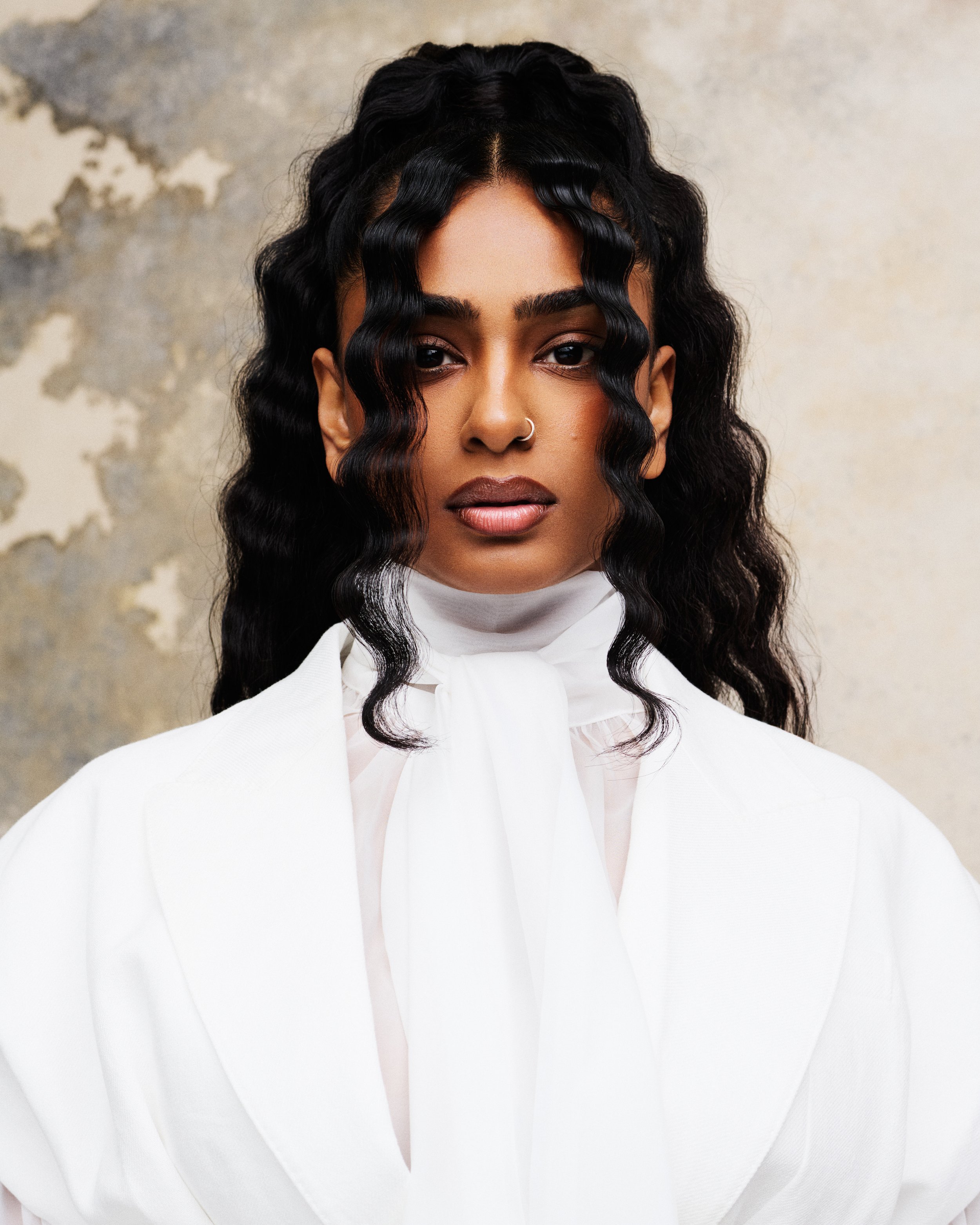 Close-up portrait of a woman with dark, wavy hair, dark eyebrows, and a nose ring, wearing a white shirt against a textured, mottled background.